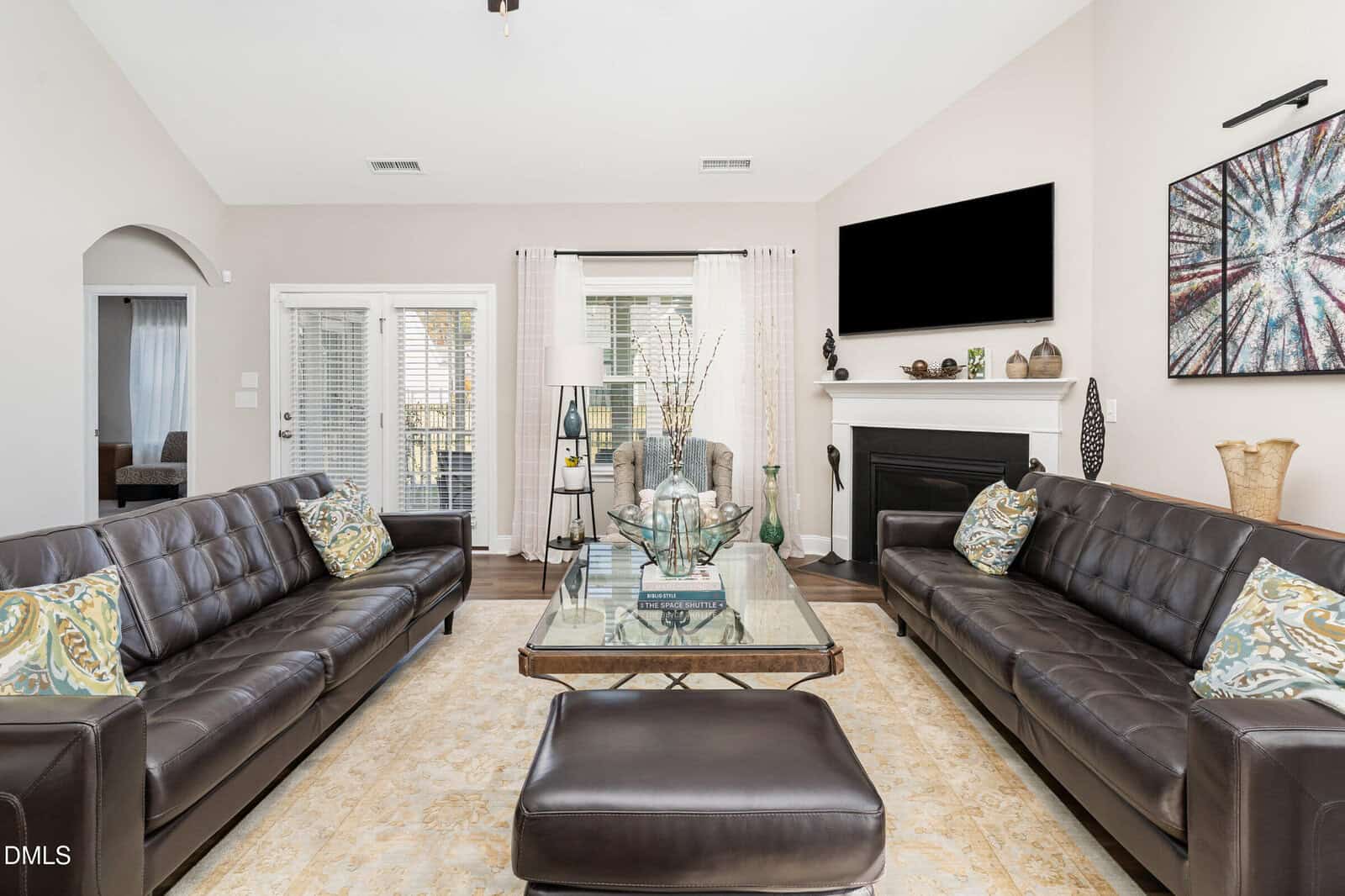 Modern living room with two dark leather sofas, a glass coffee table, a fireplace, a flat-screen TV, large windows with sheer curtains, and neutral decor. A tan area rug covers the floor.