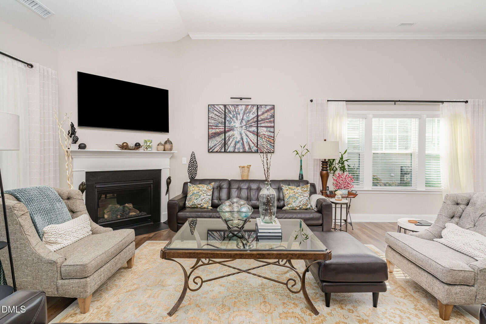 Bright living room with a black leather couch, two beige armchairs, a glass coffee table, a fireplace, wall art, and large windows. Neutral decor with patterned pillows and decorative vases.