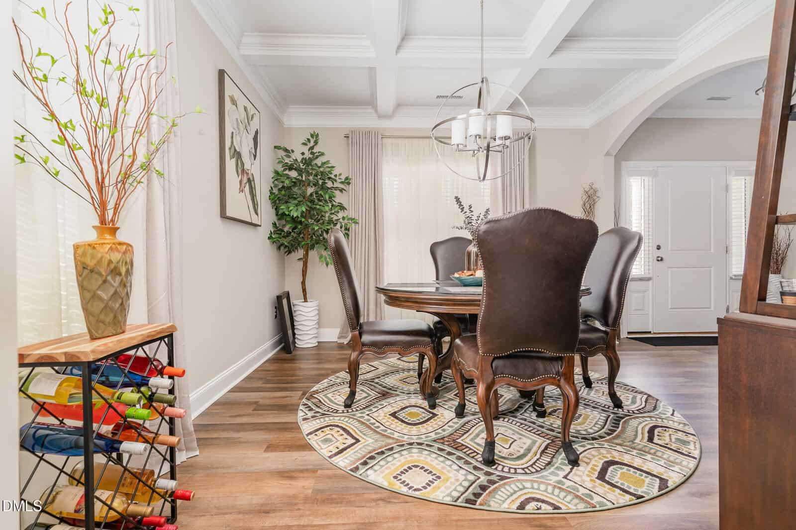 A dining room with a round table and leather chairs on a patterned rug, wine rack with bottles, potted plant, wall art, large vase, and natural light from windows with white curtains.