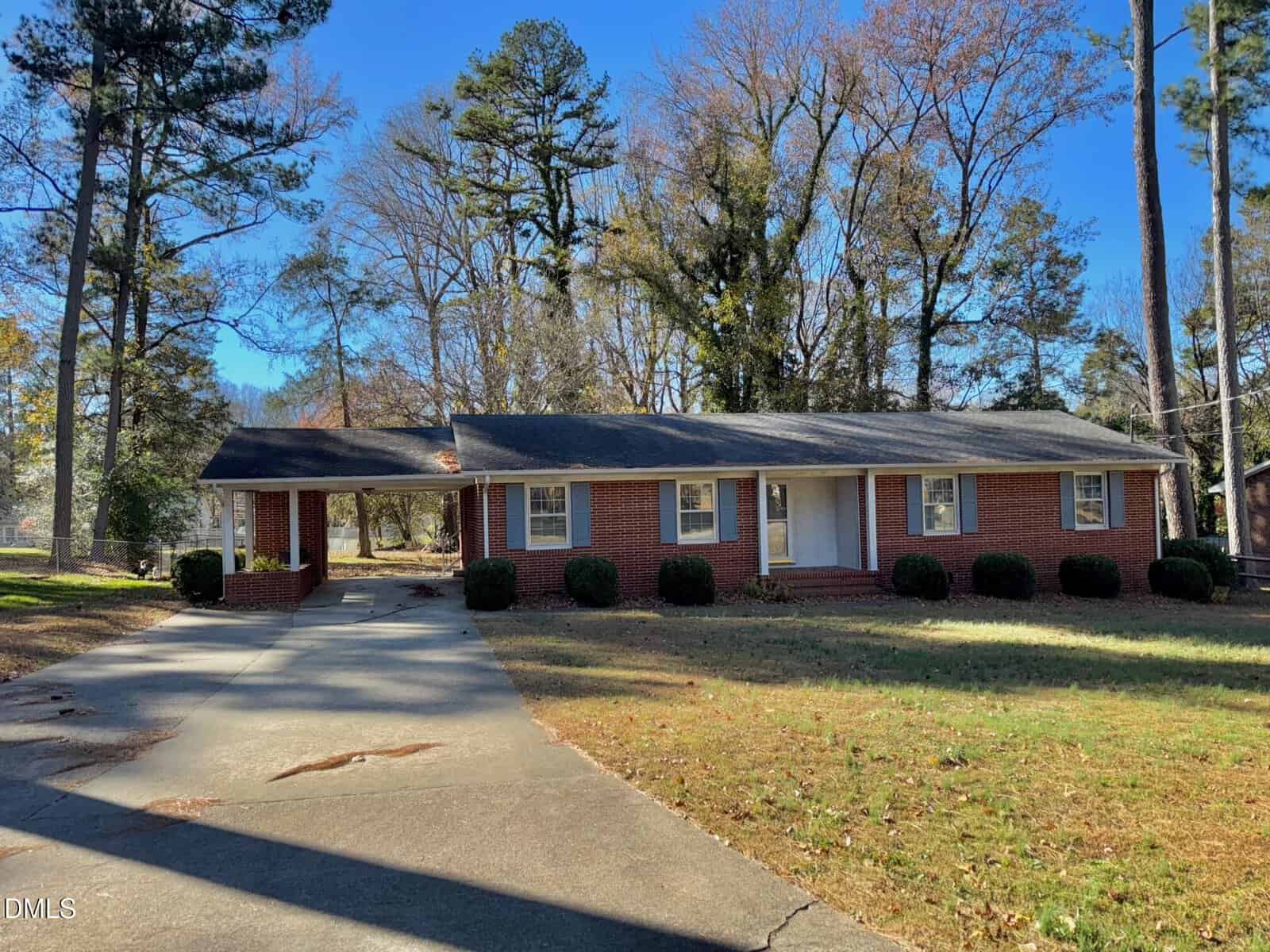 Single-story red brick house with white trim, five front windows, and a covered carport on the left. The yard features patchy grass, bushes along the front, and tall trees in the background. A curved driveway leads to the house.