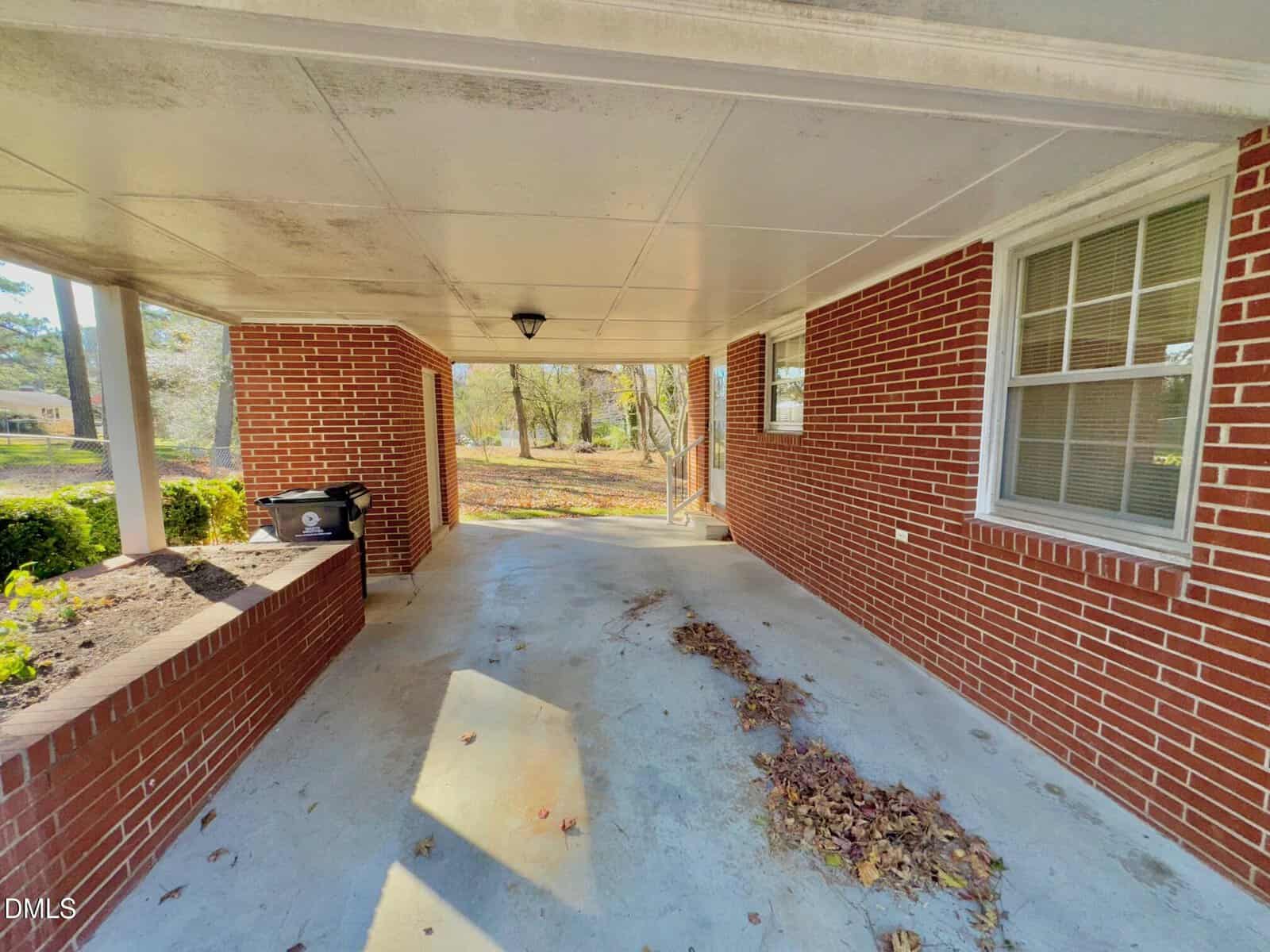A covered carport with a concrete floor, red brick walls, a window on the right, a black grill on the left, and scattered dry leaves on the ground. The area opens to a yard with trees in the background.