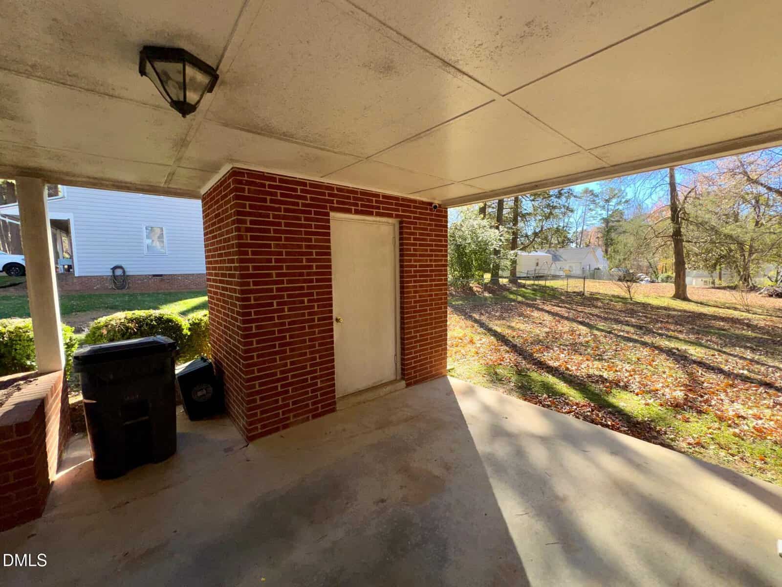 A covered patio area with a brick wall, a white door, two trash bins, and a light fixture on the ceiling. The ground is concrete, and a grassy yard with trees is visible in the background.