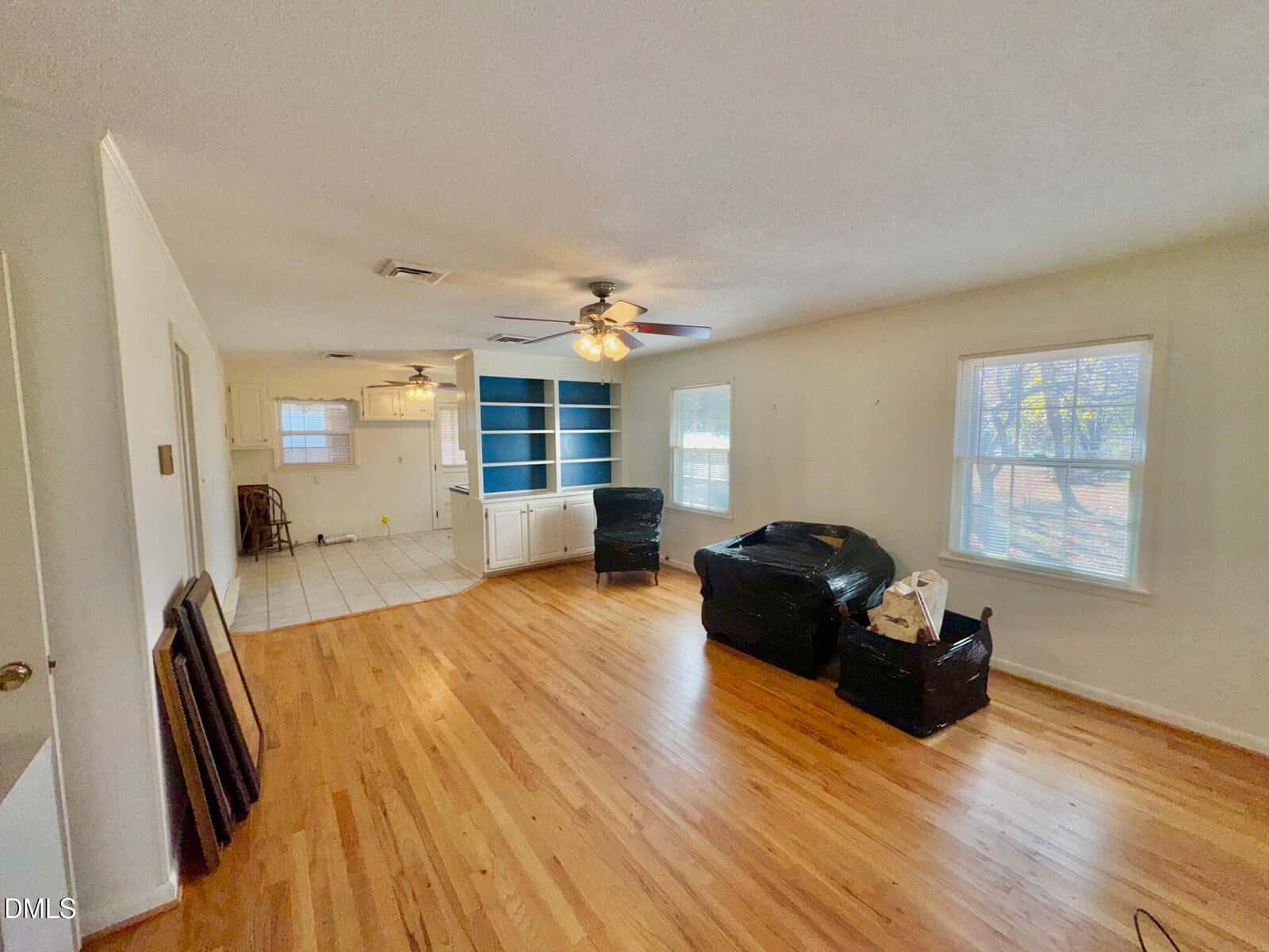 A spacious living room with hardwood floors, two ceiling fans, and large windows. The room contains some wrapped furniture and bags. In the background, there is a kitchen area and built-in shelves painted blue.
