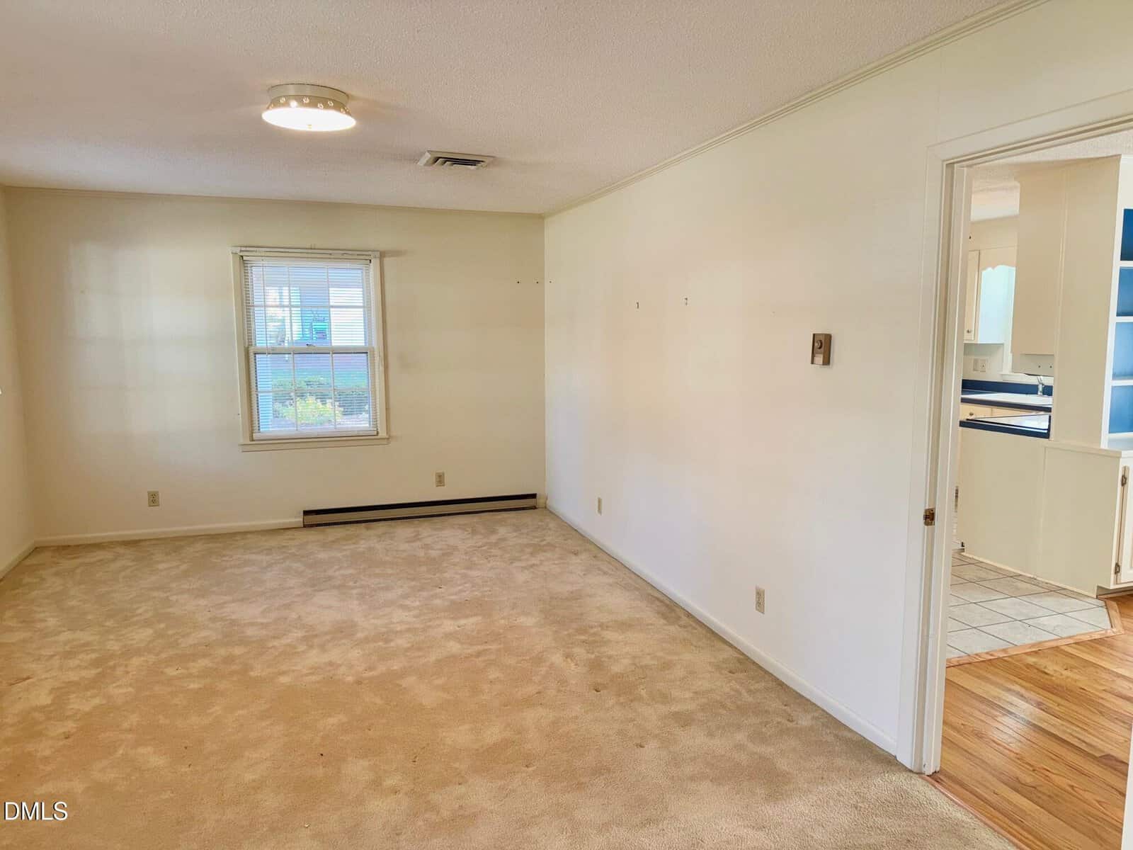 A mostly empty room with beige carpet, off-white walls, a window with blinds, a baseboard heater, and a ceiling light. An open doorway leads to a kitchen with tile flooring and visible cabinets.
