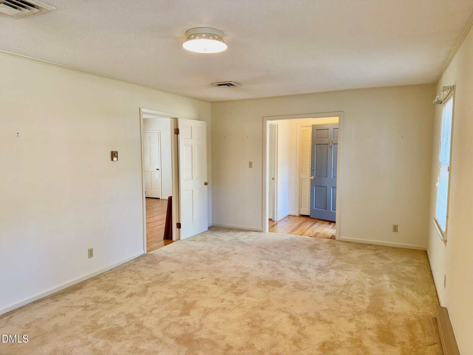 A spacious, empty room with beige carpet, white walls, two open doorways leading to other rooms, and windows letting in natural light. The ceiling has a round light fixture and there are minimal wall features.
