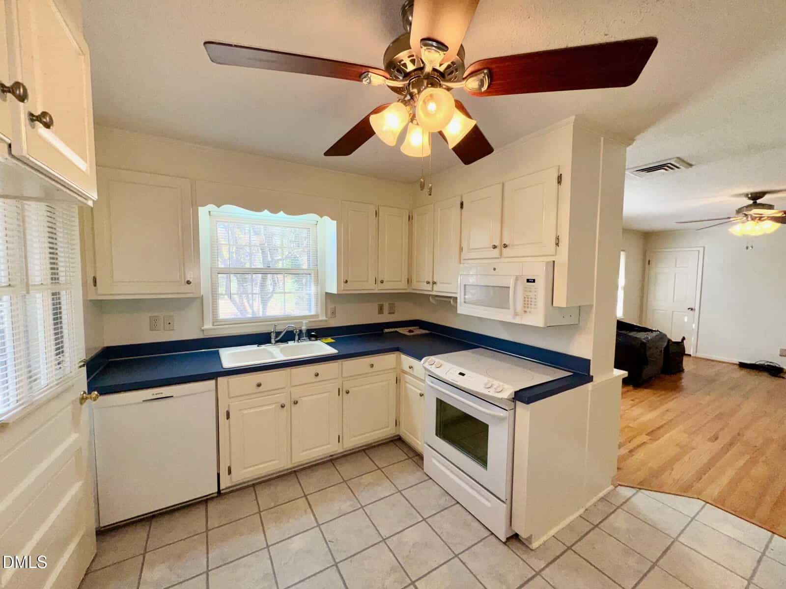 Kitchen with white cabinets, blue countertops, white appliances, tile flooring, and a ceiling fan with lights. There is a window above the sink and a view of a living area with wood flooring in the background.