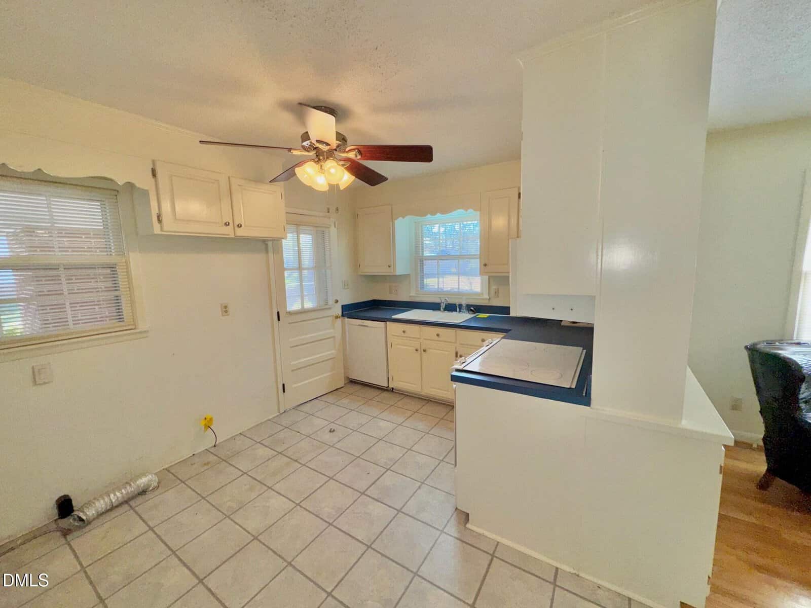 Small kitchen with white cabinets, blue countertops, a ceiling fan with lights, tiled floor, and multiple windows. An empty corner with a vent pipe suggests space for a missing appliance.
