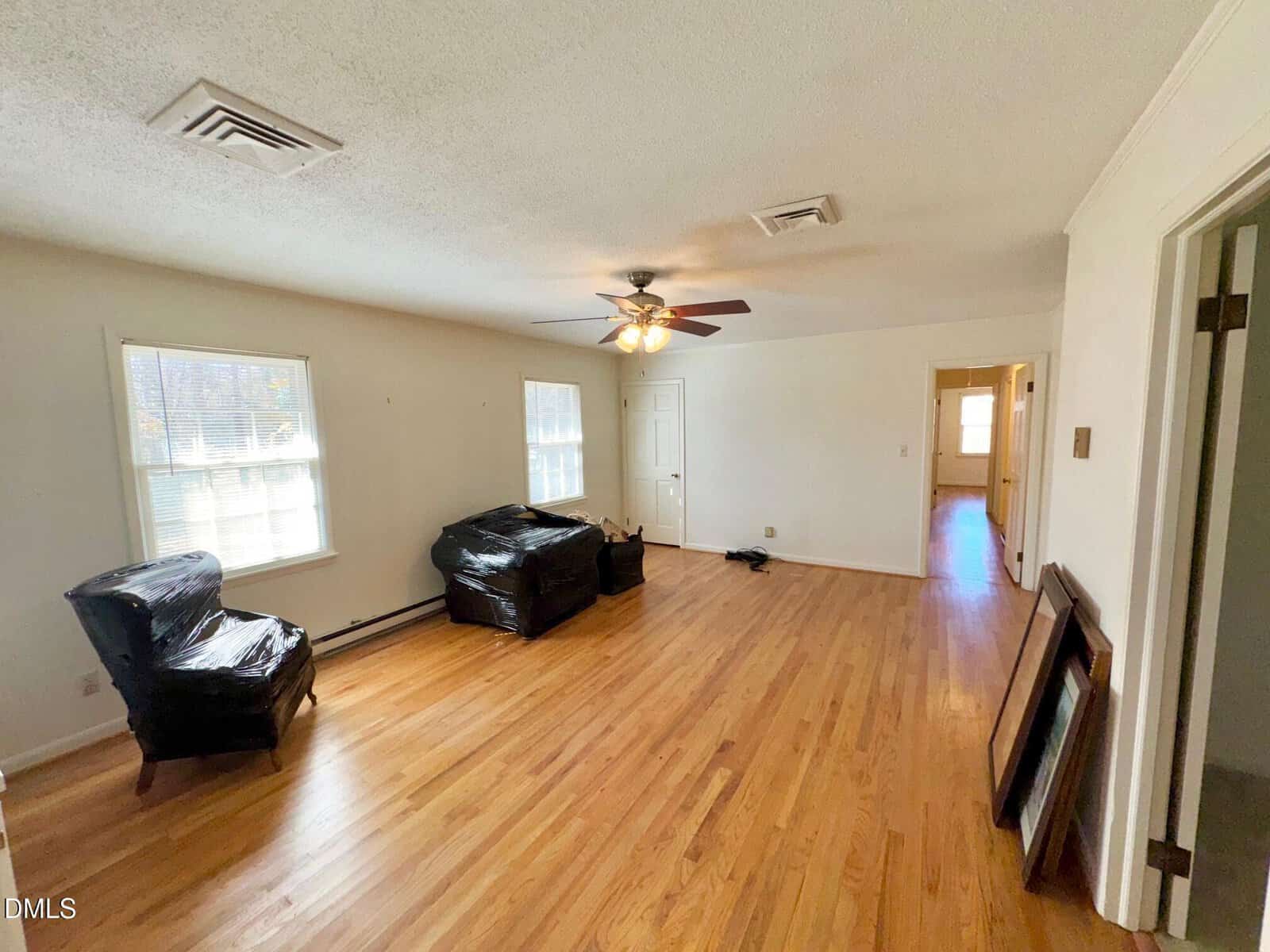 A spacious living room with hardwood floors, two windows, a ceiling fan with lights, an armchair and a loveseat covered in plastic, and an open doorway leading to another room.