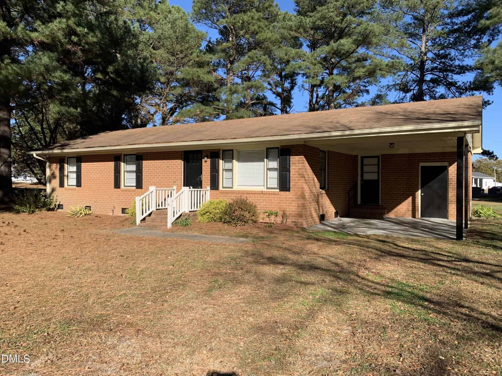 A charming one-story brick house at 41 Tant Road, Zebulon, features a gabled roof, white trim, and a small front porch with railings. The yard is blanketed in pine needles and surrounded by tall trees, with an attached carport on the right.