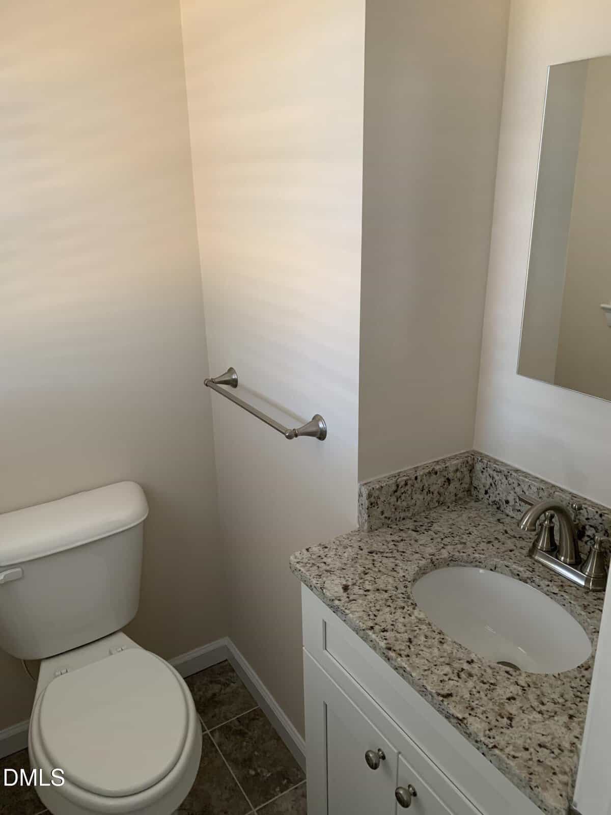 Small bathroom with a white toilet, granite countertop sink with white cabinet, silver faucet, wall-mounted mirror, and a silver towel bar on a light-colored wall. Brown tile flooring is visible.