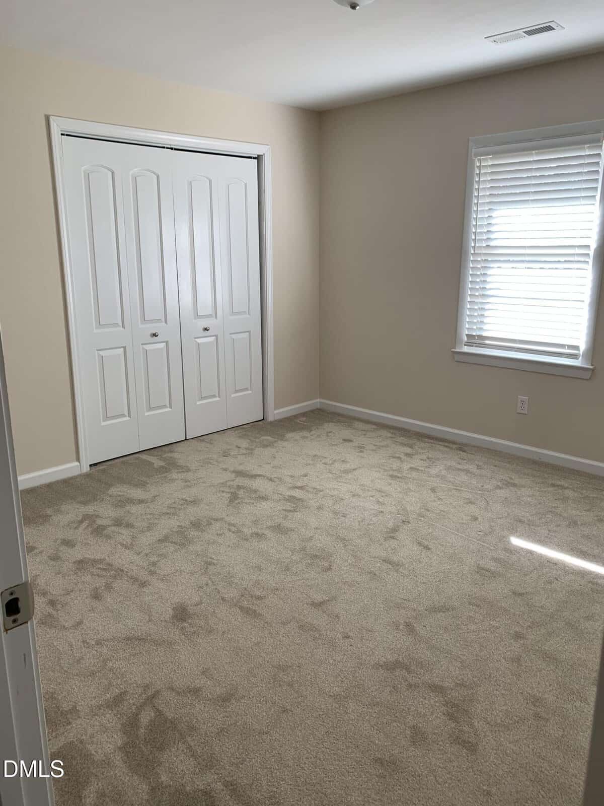An empty, carpeted bedroom with beige walls, a window with white blinds, and a double-door closet. Sunlight enters through the window, creating a bright and clean appearance.