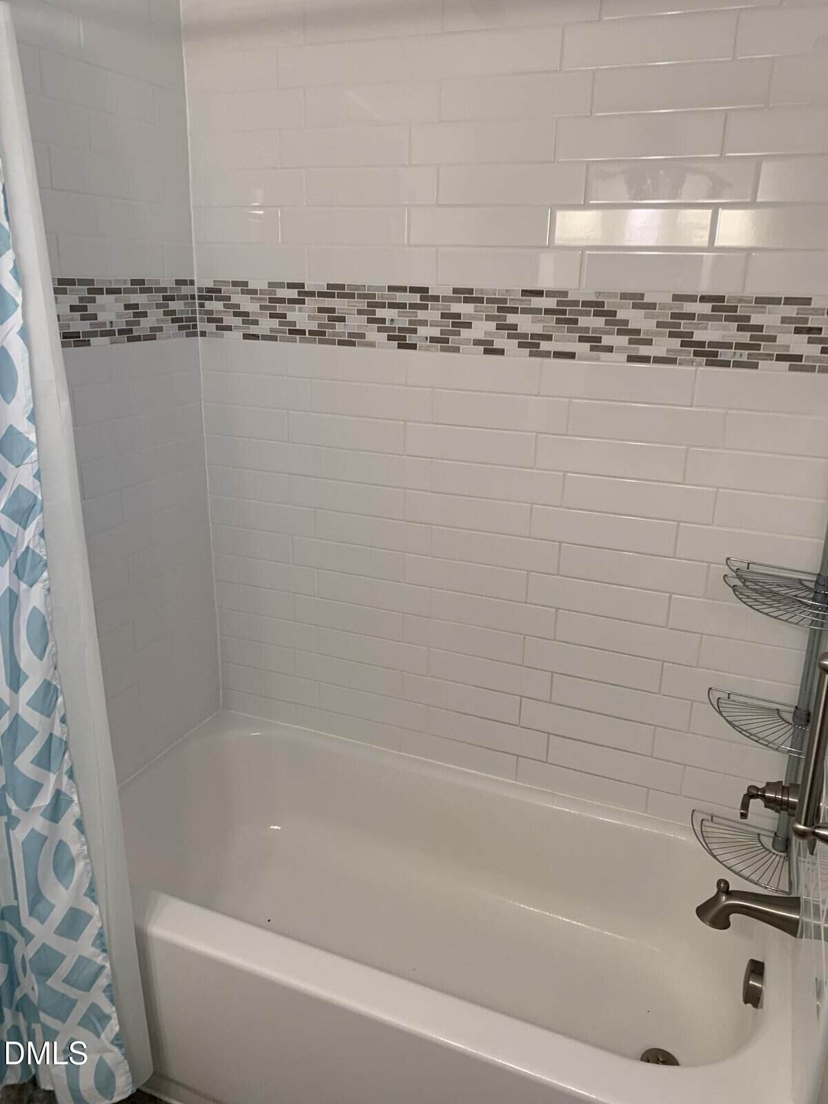 A white bathtub with white subway tile walls, accented by a horizontal band of small gray and brown tiles, a chrome shower fixture, a corner metal shelf, and a blue patterned shower curtain.