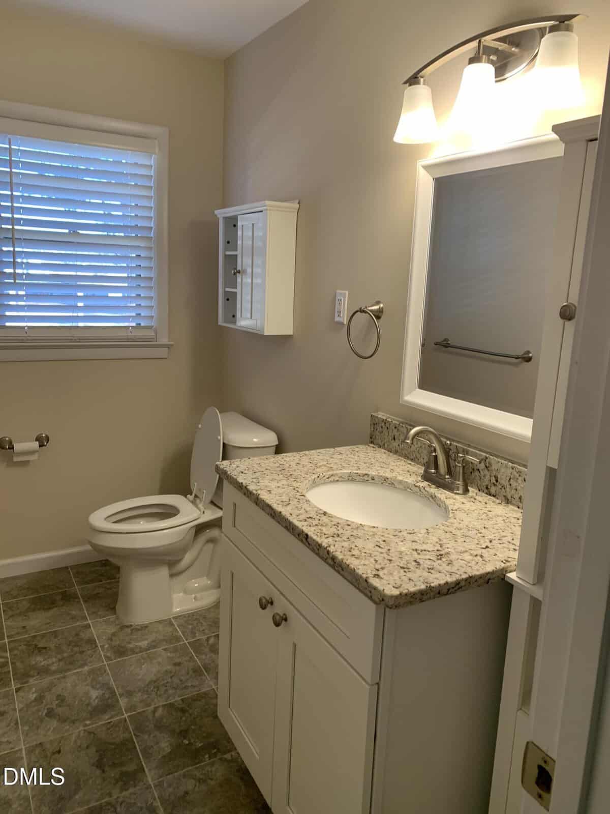 A bathroom with a granite countertop vanity, white sink, wall-mounted mirror with lights, toilet, wall cabinet, towel bar, and a window with blinds. The floor is tiled in gray.