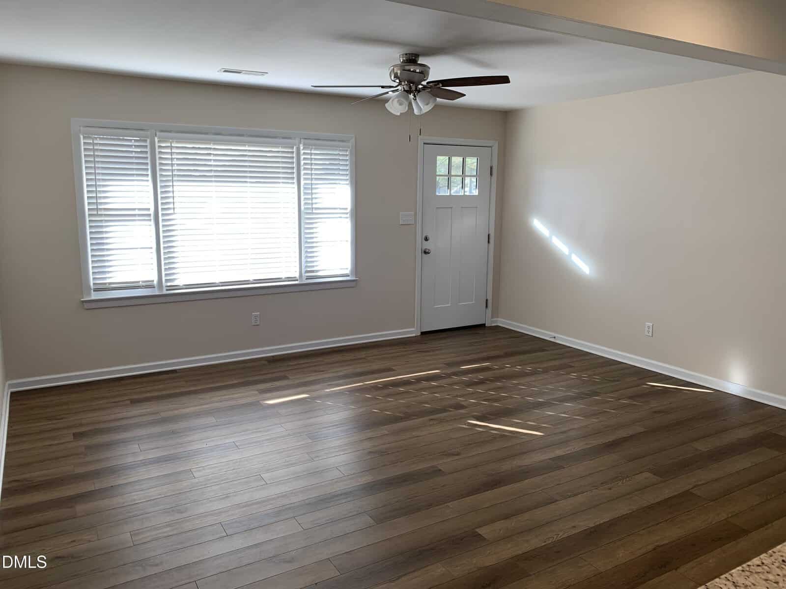 A bright, empty room with light beige walls, wood-style flooring, a white front door, large windows with blinds, and a ceiling fan with lights. Sunlight streams in, casting shadows on the floor.