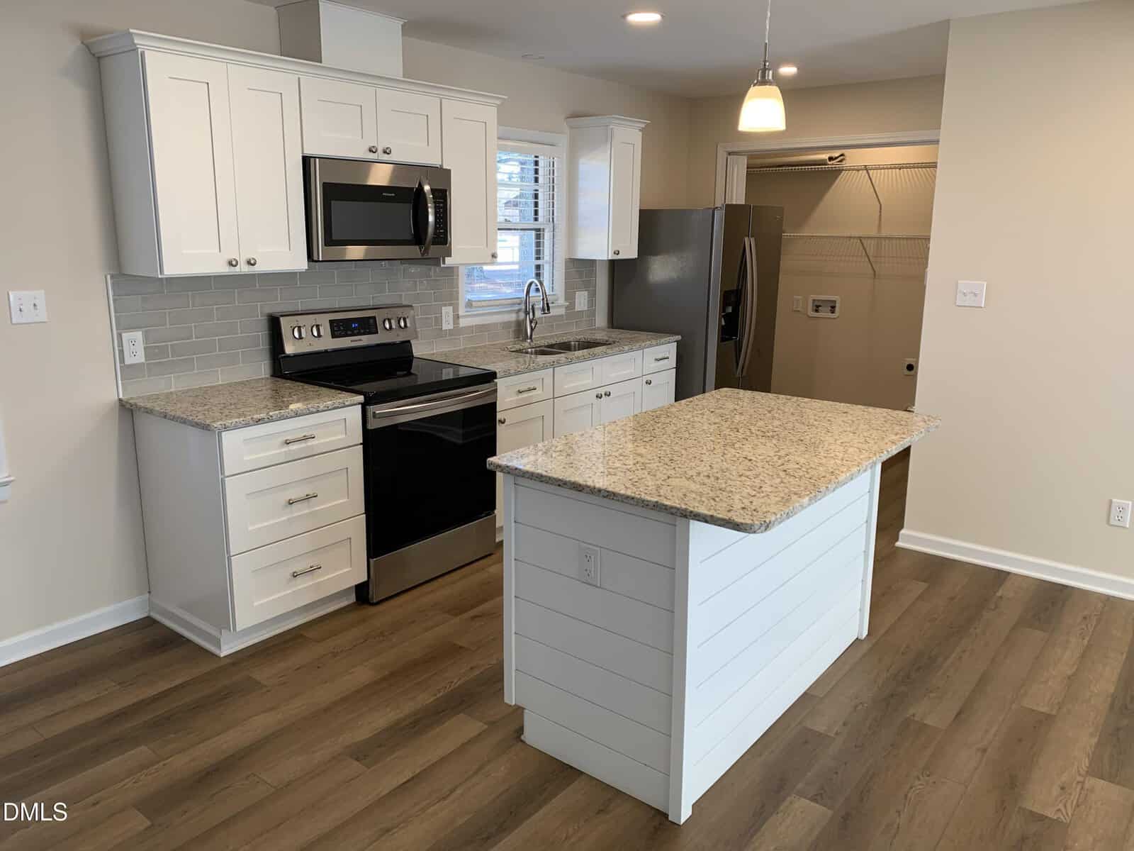 Modern kitchen with white cabinets, stainless steel appliances, a granite island, subway tile backsplash, and wood-look flooring. Refrigerator and pantry visible in the background; pendant light hangs above the island.