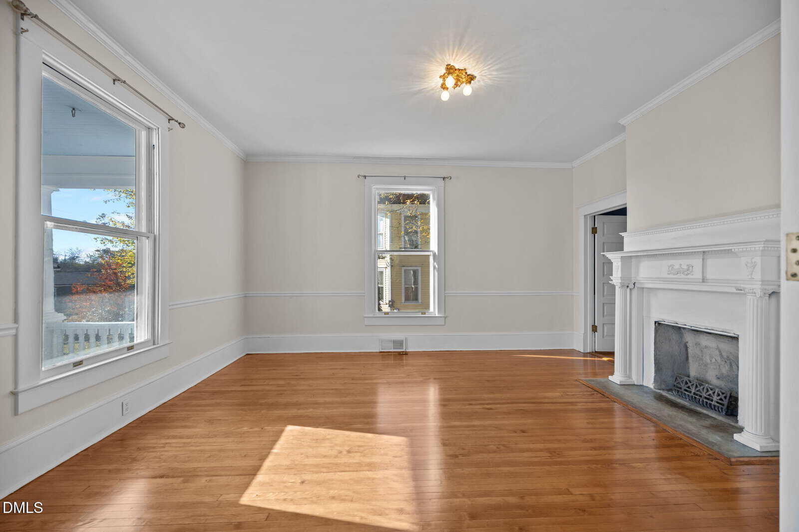 Bright, empty living room with light beige walls, hardwood floors, a white fireplace, and large windows letting in natural light. A simple ceiling light fixture is at the center of the ceiling.