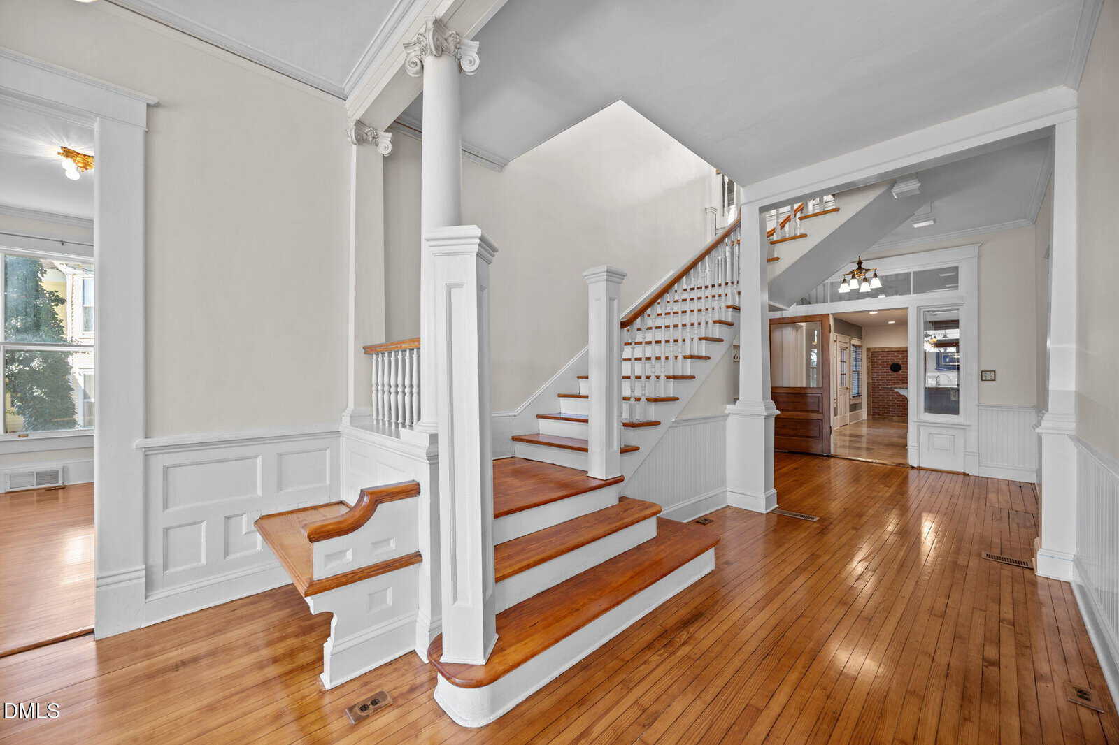 Bright interior of a house with polished wood floors, white wainscoting and columns, a wooden staircase with white balusters, and natural light coming from large windows and open doorways.