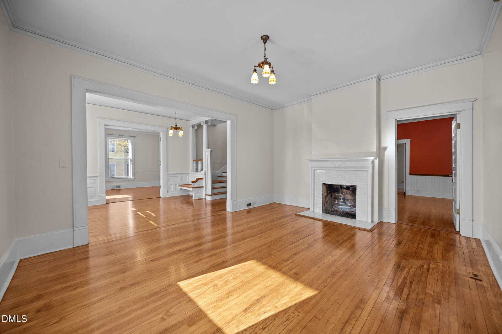 A spacious living room with light wood floors, cream walls, a white fireplace, and vintage ceiling lights. Large doorways connect to adjoining rooms with similar flooring and white trim, and sunlight streams through the windows.