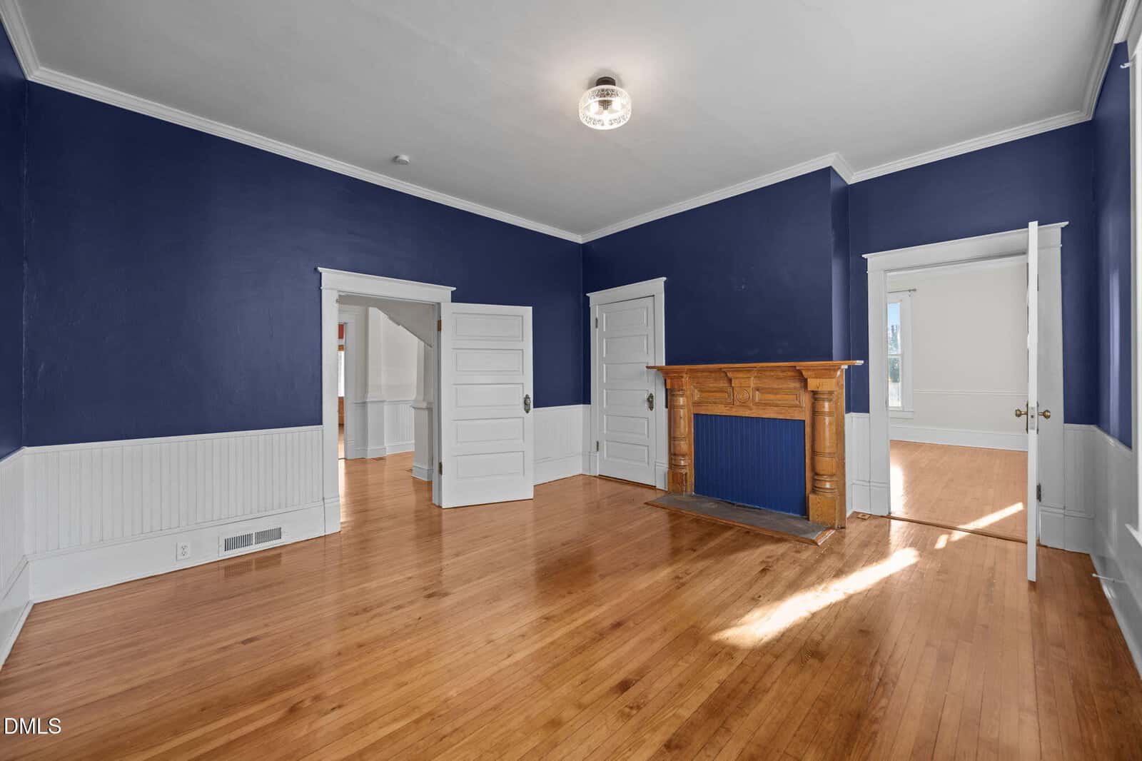 A spacious room with dark blue walls, white trim, and hardwood floors. There is a wooden fireplace with a blue accent, several white doors, and natural light from windows in an adjacent room.
