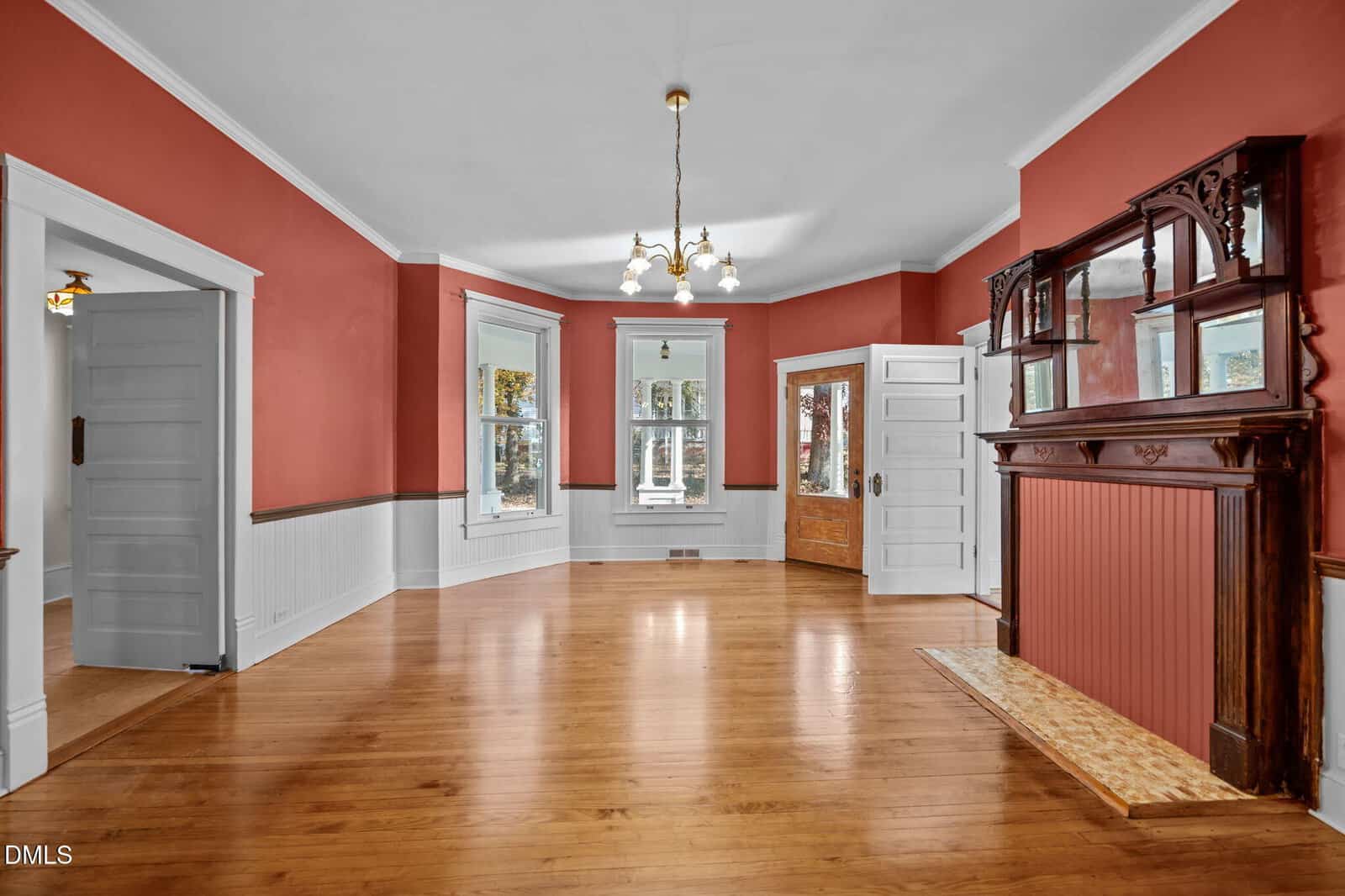 A spacious room with red and white walls, hardwood floors, large windows, a wooden door with glass panel, brass chandelier, and an ornate wood fireplace with mirror and mantle.