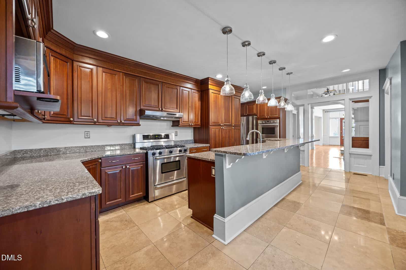 Modern kitchen with dark wood cabinets, stainless steel appliances, granite countertops, a large island with pendant lights, and beige tiled floor. Wide view shows open hallway and natural light from adjacent rooms.