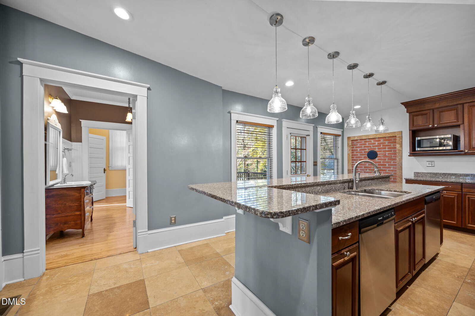 Modern kitchen with granite countertops, stainless steel appliances, pendant lights, wood cabinets, and a brick accent wall. Large windows and an open doorway lead to another room with wood flooring and a vanity.