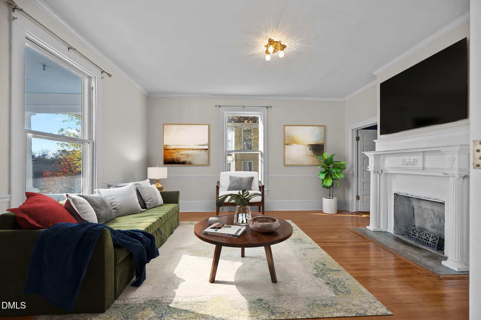 Bright living room with a green sofa, round coffee table, and white armchair. Large windows, two abstract paintings, potted plant, and a white fireplace with a TV above it. Wood floors and neutral walls complete the space.