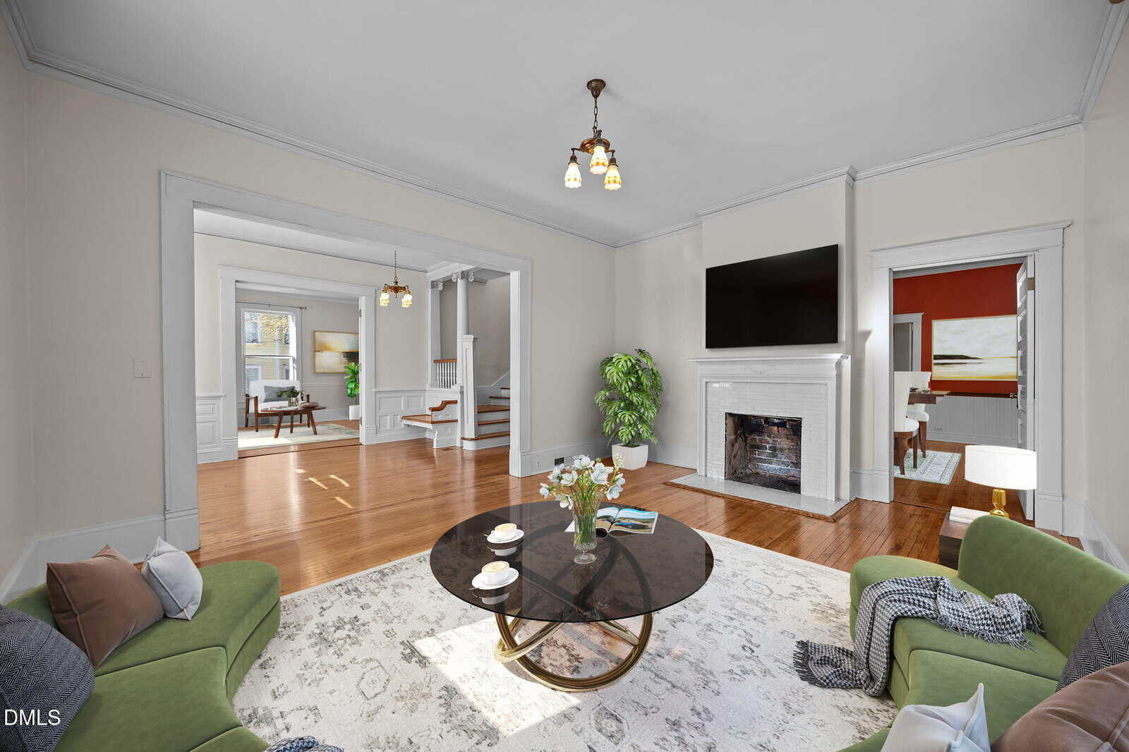 Bright living room with green sofas, round glass coffee table, and a white fireplace beneath a mounted TV. Hardwood floors, large area rug, and open doorways lead to dining area and staircase. Plant and wall art add decor.