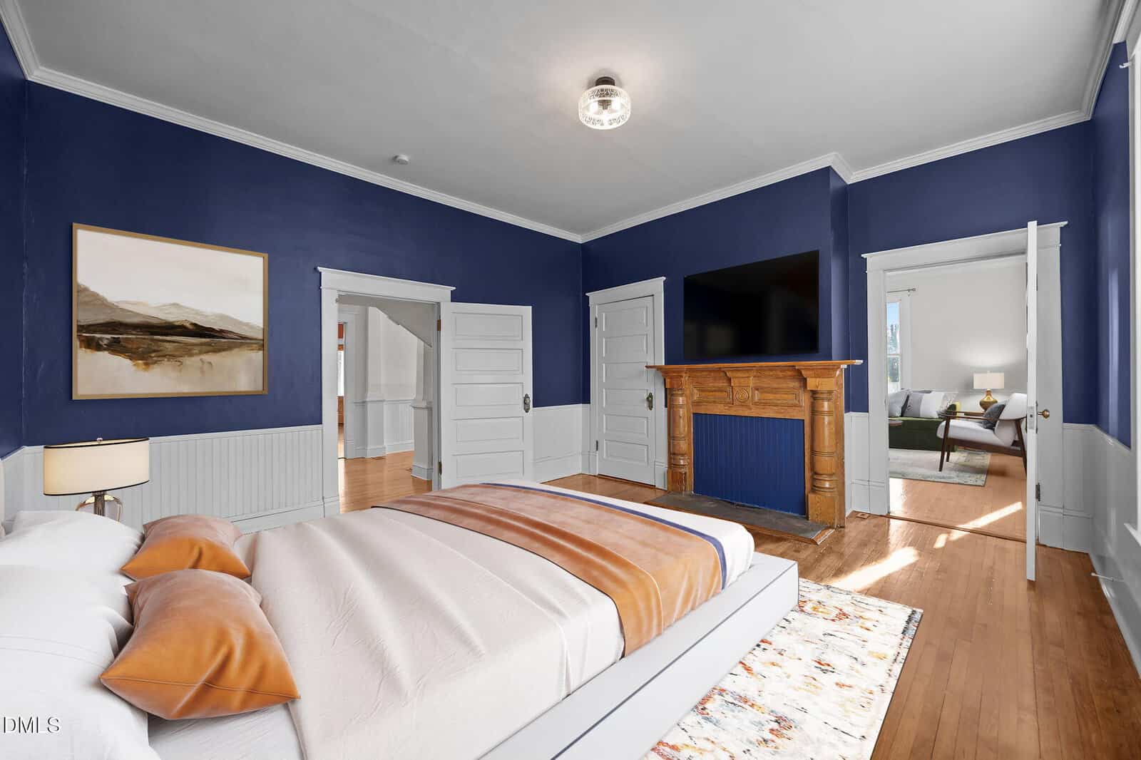 A bedroom with navy blue walls, a white ceiling, and wood floors features a modern bed with tan blankets and pillows, an orange brick fireplace, large wall TV, and multiple white doors. Sunlight enters from an adjoining room.