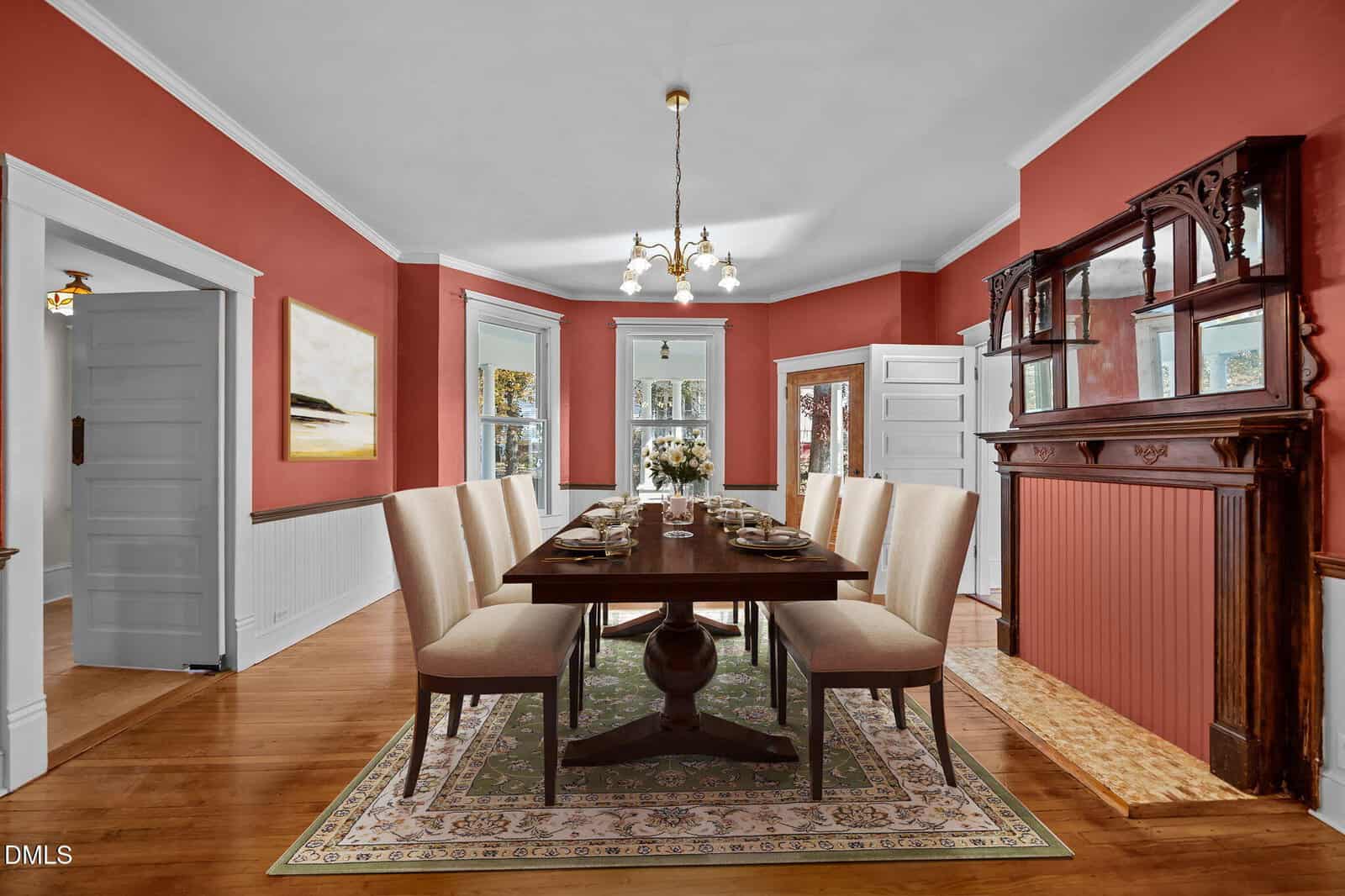 A formal dining room with rich red walls, a wooden table set for eight, cream upholstered chairs, a green patterned rug, a decorative fireplace, and large windows letting in natural light.