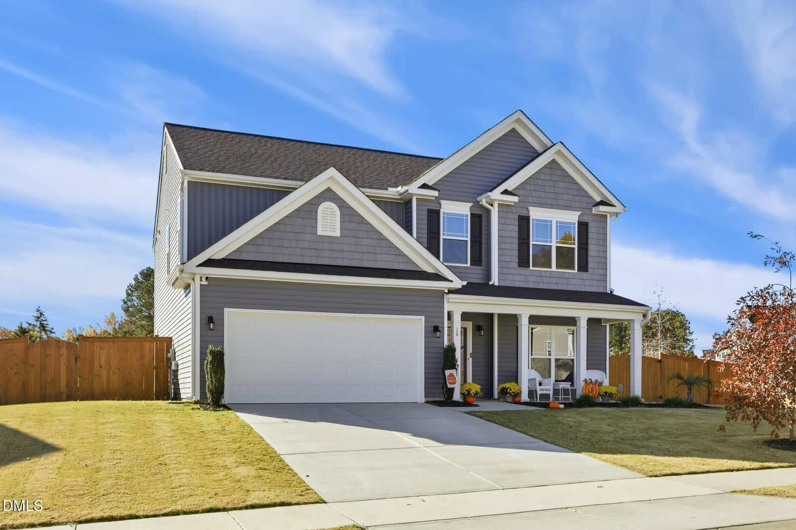 Two-story house with gray siding, white trim, and a double garage. The front porch has white chairs and potted plants, and the yard is neatly manicured with a wooden fence surrounding the backyard.