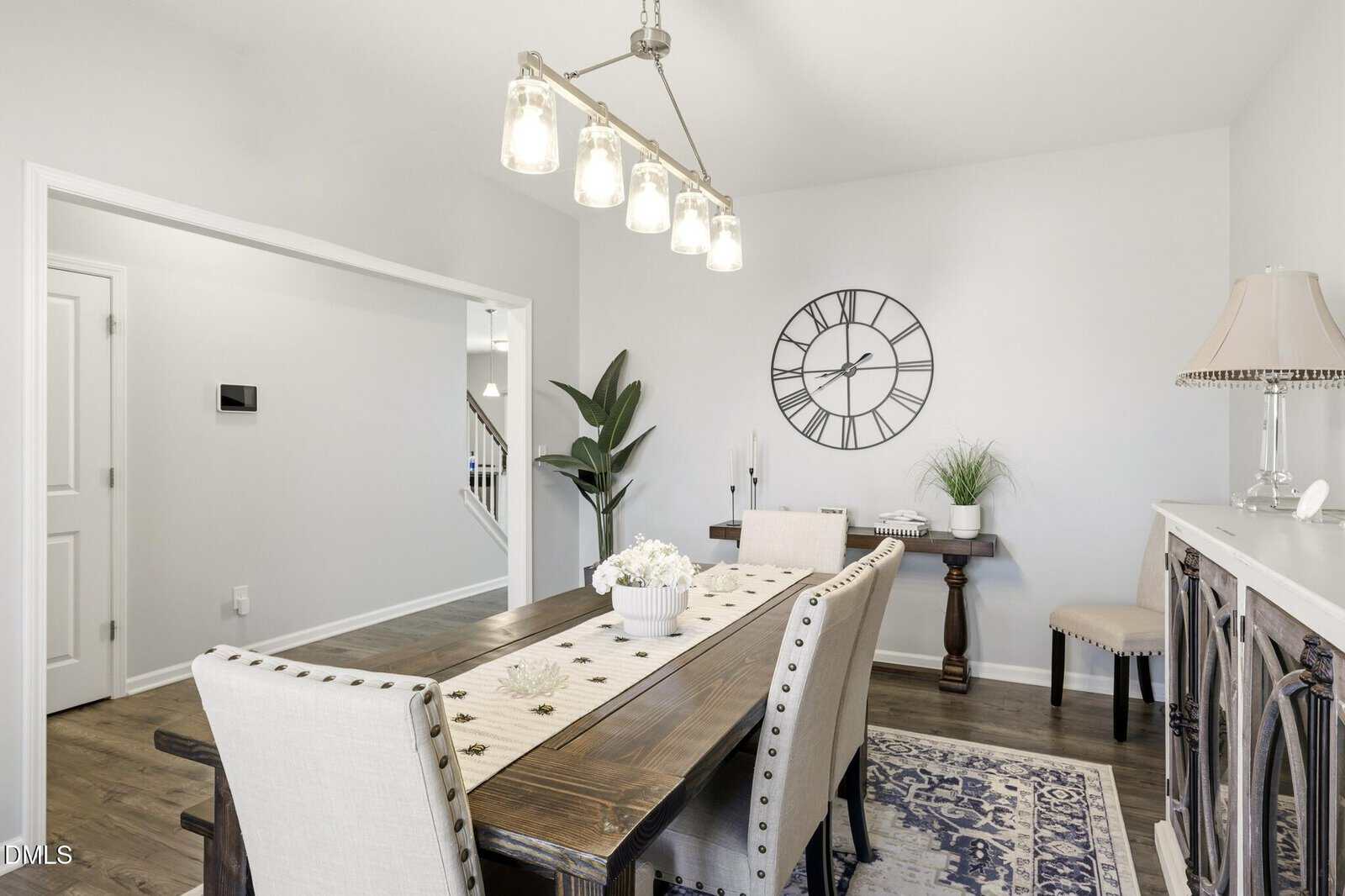 Bright dining room with a wooden table, upholstered chairs, a table runner, a plant, large wall clock, sideboard, lamp, and decorative accents on hardwood flooring. Natural light fills the space.