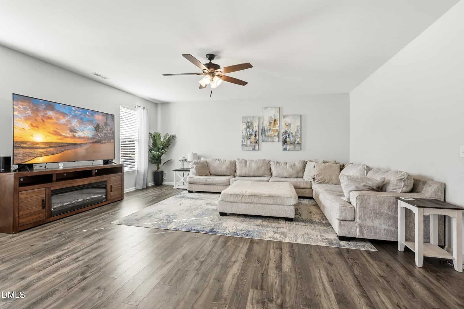 Spacious living room with a large beige sectional sofa, a wooden TV stand with a flat-screen TV displaying a beach scene, a ceiling fan, abstract wall art, and wood flooring. Light streams in from a window with white curtains.