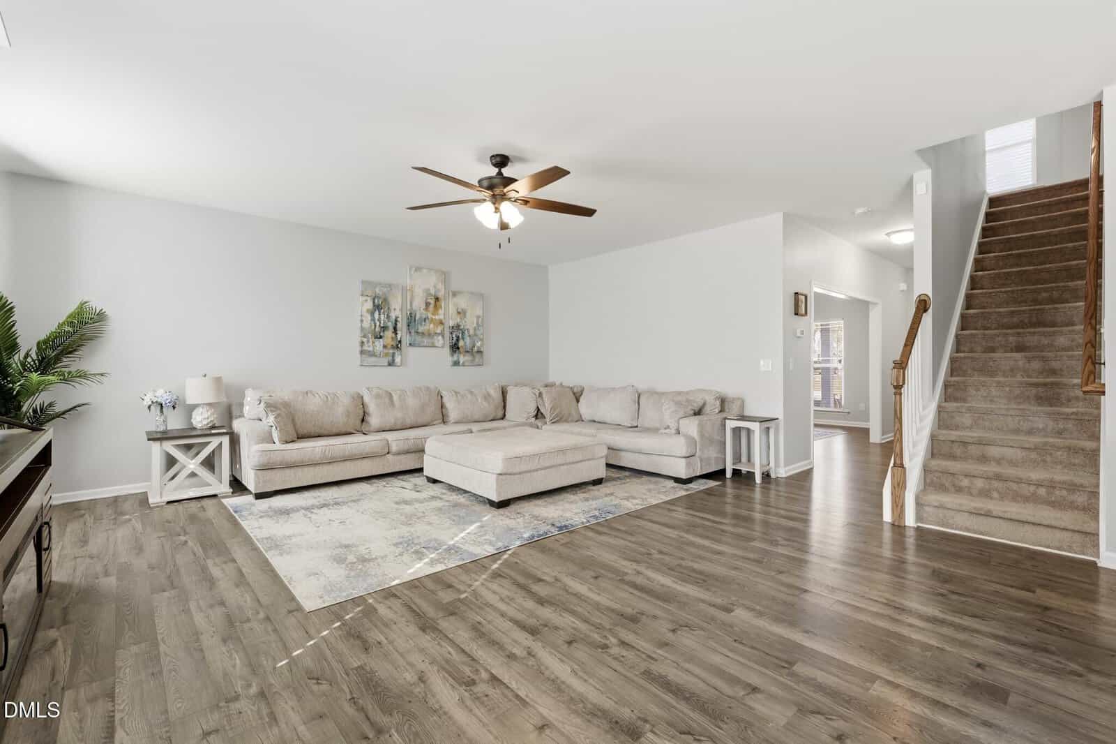 Spacious living room with a large beige sectional sofa, abstract art on the wall, a ceiling fan, wood flooring, a potted plant, and carpeted stairs leading upward on the right.