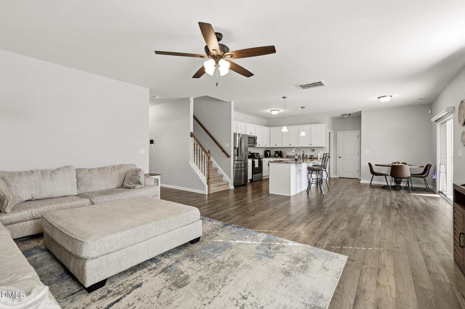 Open-concept living space with a beige sectional, ceiling fan, kitchen with stainless steel appliances, dining area with round table, wooden floors, and stairway leading upstairs. Ample natural light throughout the room.