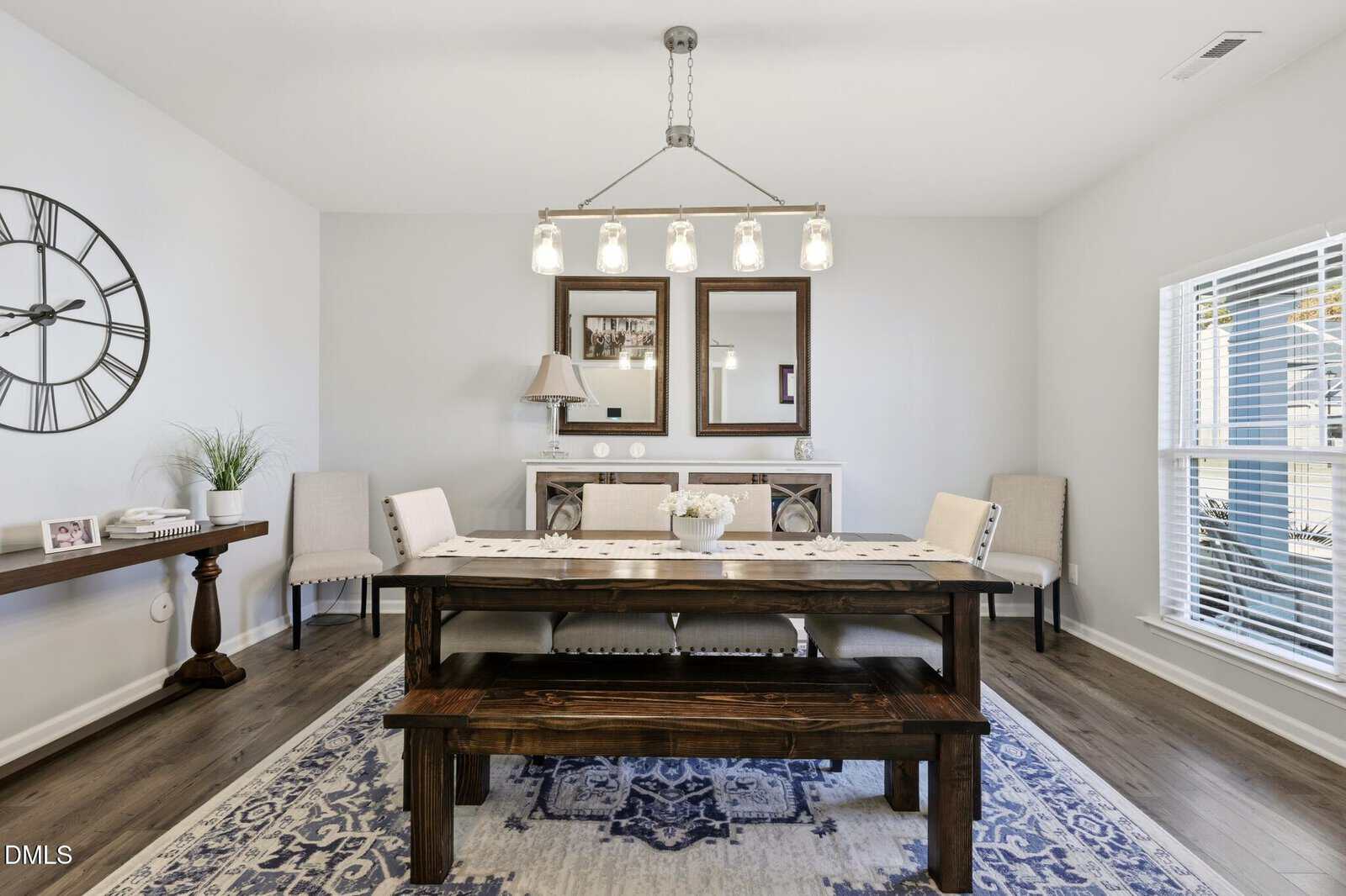 A modern dining room with a dark wood table, matching bench, four upholstered chairs, a large wall clock, sideboard with decor, three mirrors, and a window with white blinds letting in natural light.