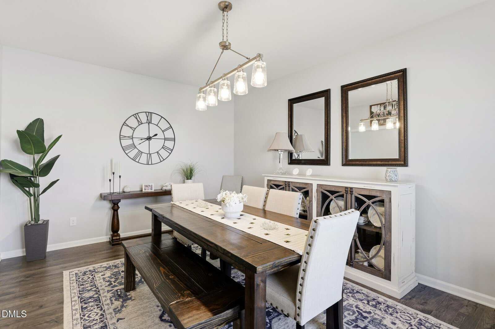 A modern dining room with a wooden table, beige upholstered chairs, and a bench. Two mirrors and a lamp are on a sideboard, a clock and decor on a wall console, and a large potted plant is in the corner.