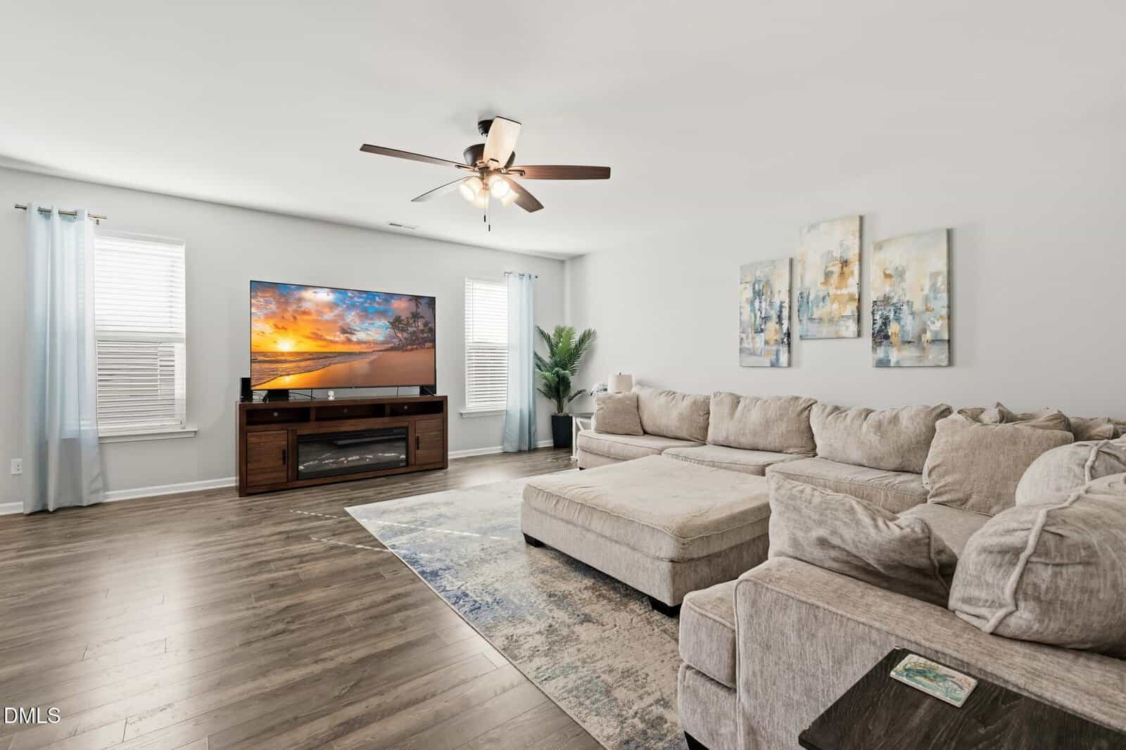Spacious living room with light walls, wood flooring, a large beige sectional sofa, a TV displaying a beach scene, a ceiling fan, wall art, and windows with sheer blue curtains.
