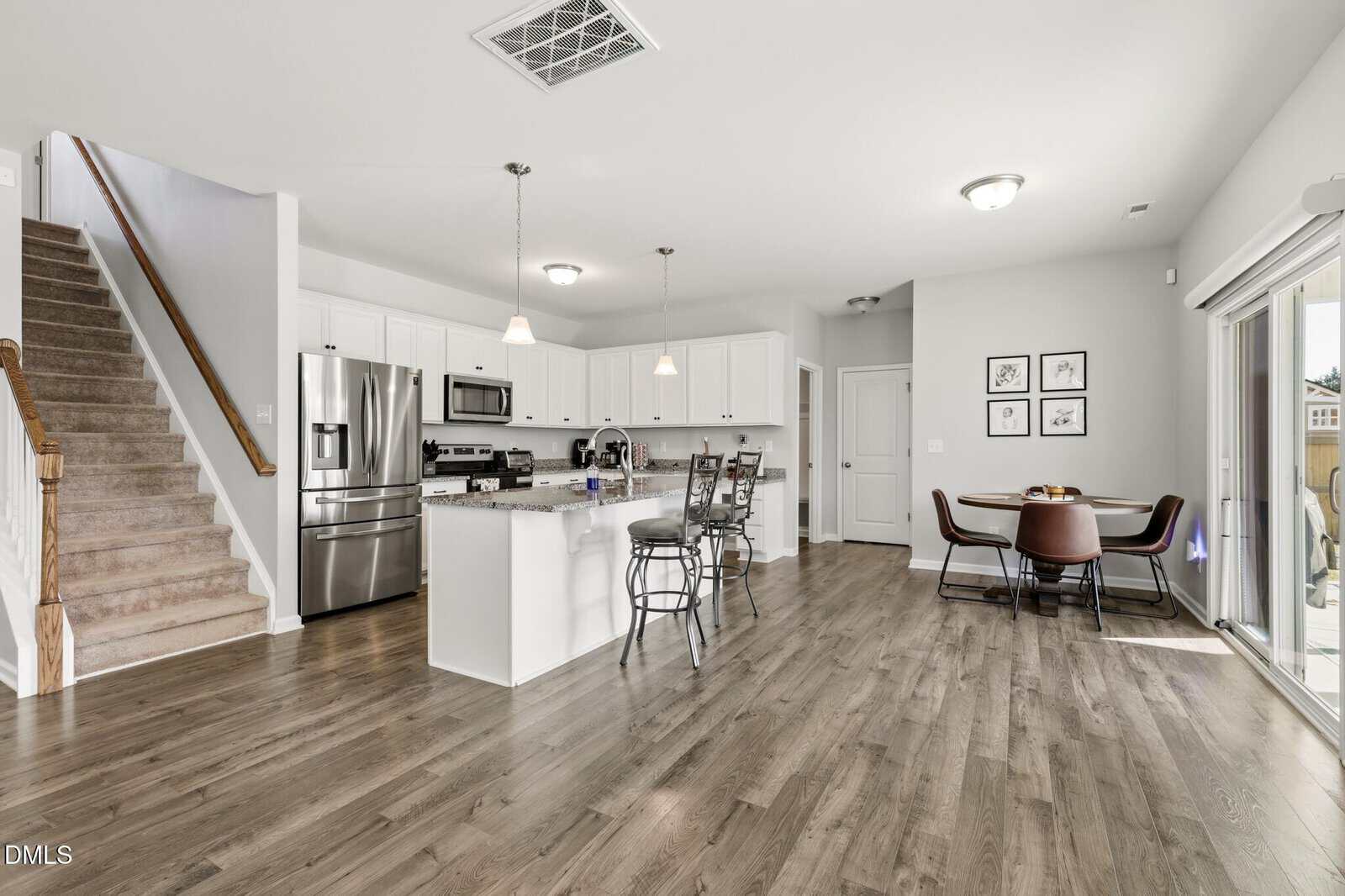 Modern open kitchen with white cabinets, stainless steel appliances, island with stools, adjacent dining area with table and chairs, stairs to the left, sliding glass door to the right, and wood flooring throughout.