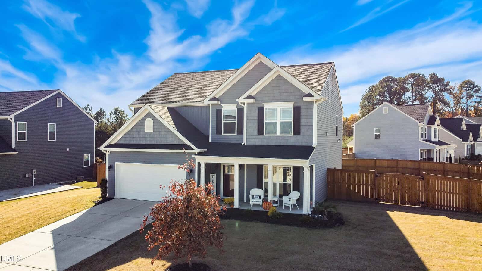 A two-story gray house with white trim, a double garage, and a front porch with two chairs. The house is surrounded by a lawn, a small tree, and is set in a suburban neighborhood under a blue sky.