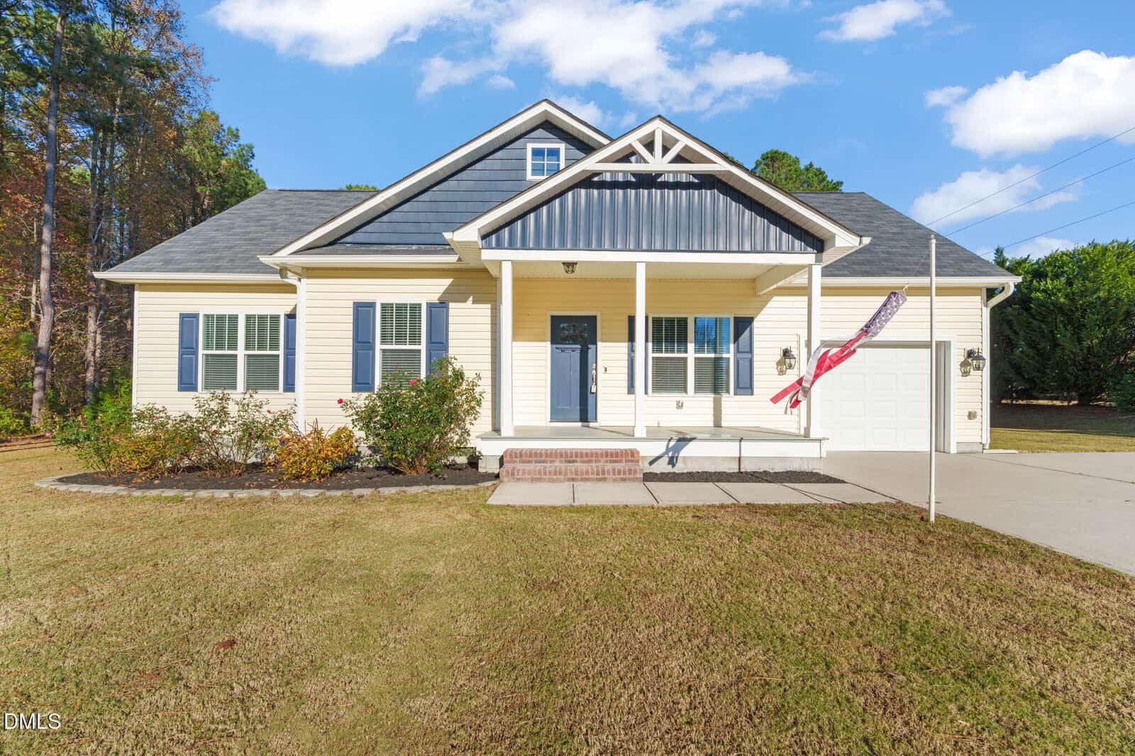 Charming single-story house at 10 Chris Ridge Way, Louisburg, featuring light yellow siding, a black roof, blue shutters, and a welcoming front porch with white columns. A brick step leads up to the entrance, and there’s an attached single-car garage. An American flag hangs proudly near the driveway. The spacious yard is grassy with well-maintained shrubs.