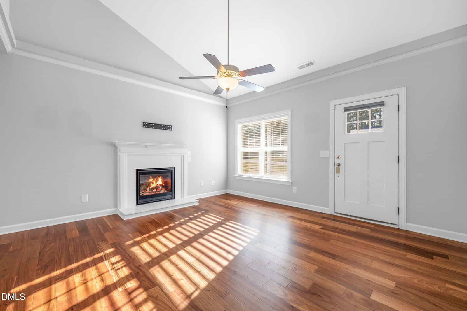 A bright, empty room with hardwood floors, a white fireplace with a lit fire, a ceiling fan with a light, a window with blinds, and a white door. Sunlight creates shadows on the floor. Walls are painted light gray.