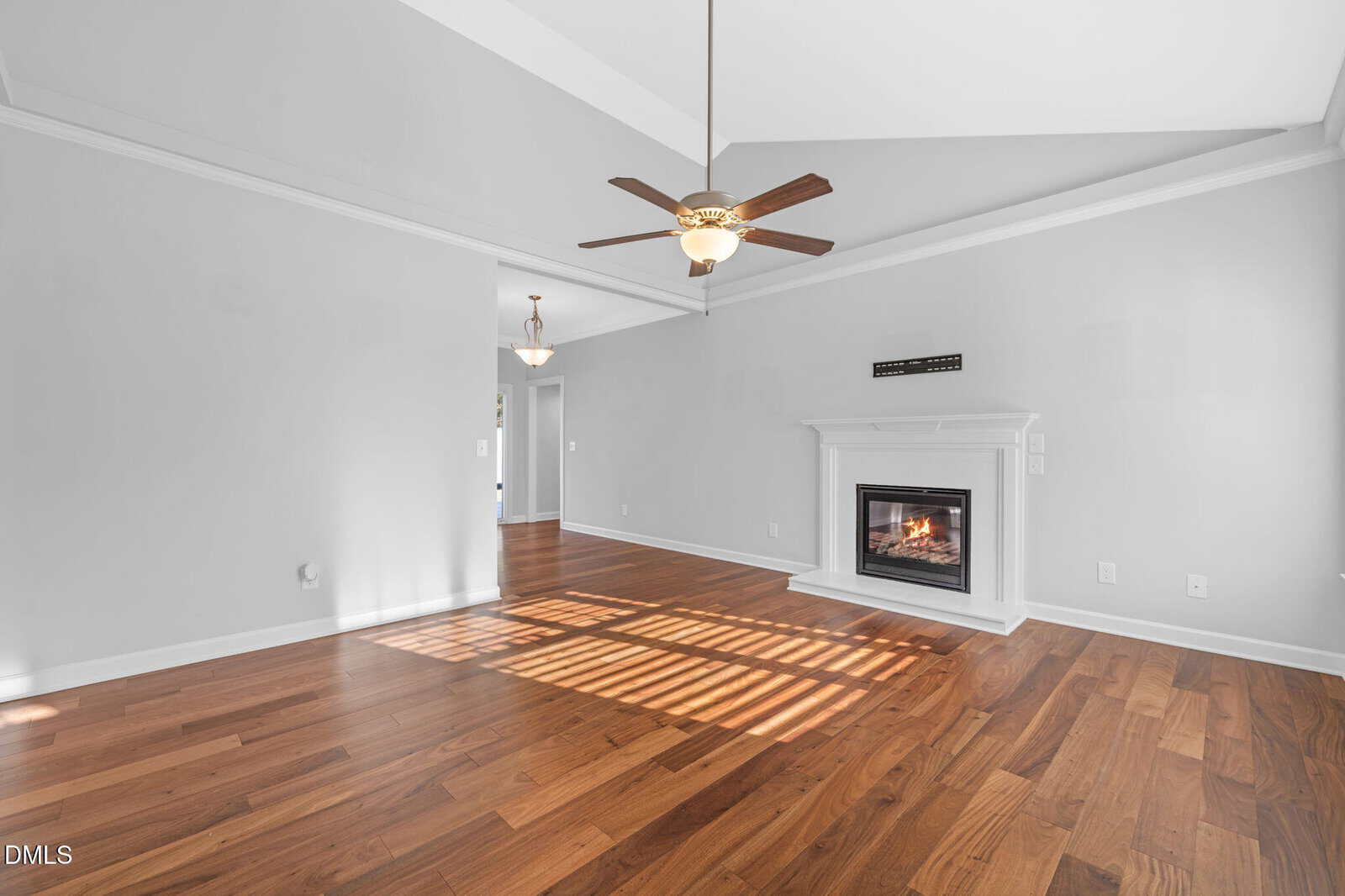 A spacious living room with light gray walls, a vaulted ceiling, ceiling fan, wood flooring, a white fireplace on the far wall, and sunlight casting shadows through a window. An open doorway leads to another room.
