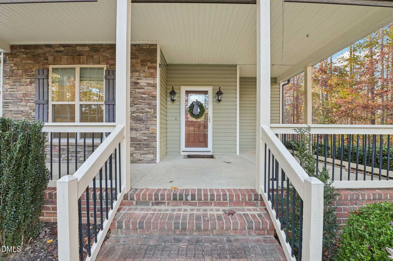 A front porch with brick steps, white railings, and beige siding. The wooden front door has a wreath, and two lantern-style lights are mounted on either side. A window with stone accents is on the left. Trees with autumn leaves are visible.
