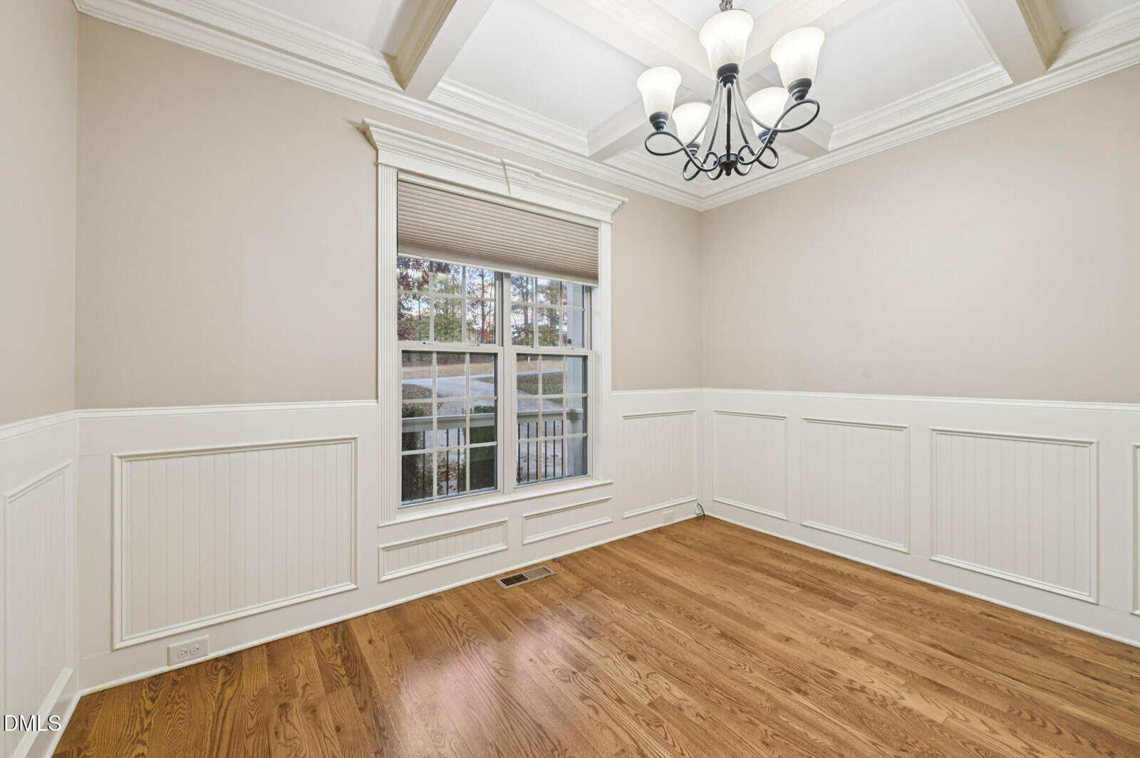 Empty room with light beige walls, white wainscoting, wooden floor, large window with blinds, and a black chandelier hanging from a coffered ceiling. Trees are visible outside the window.