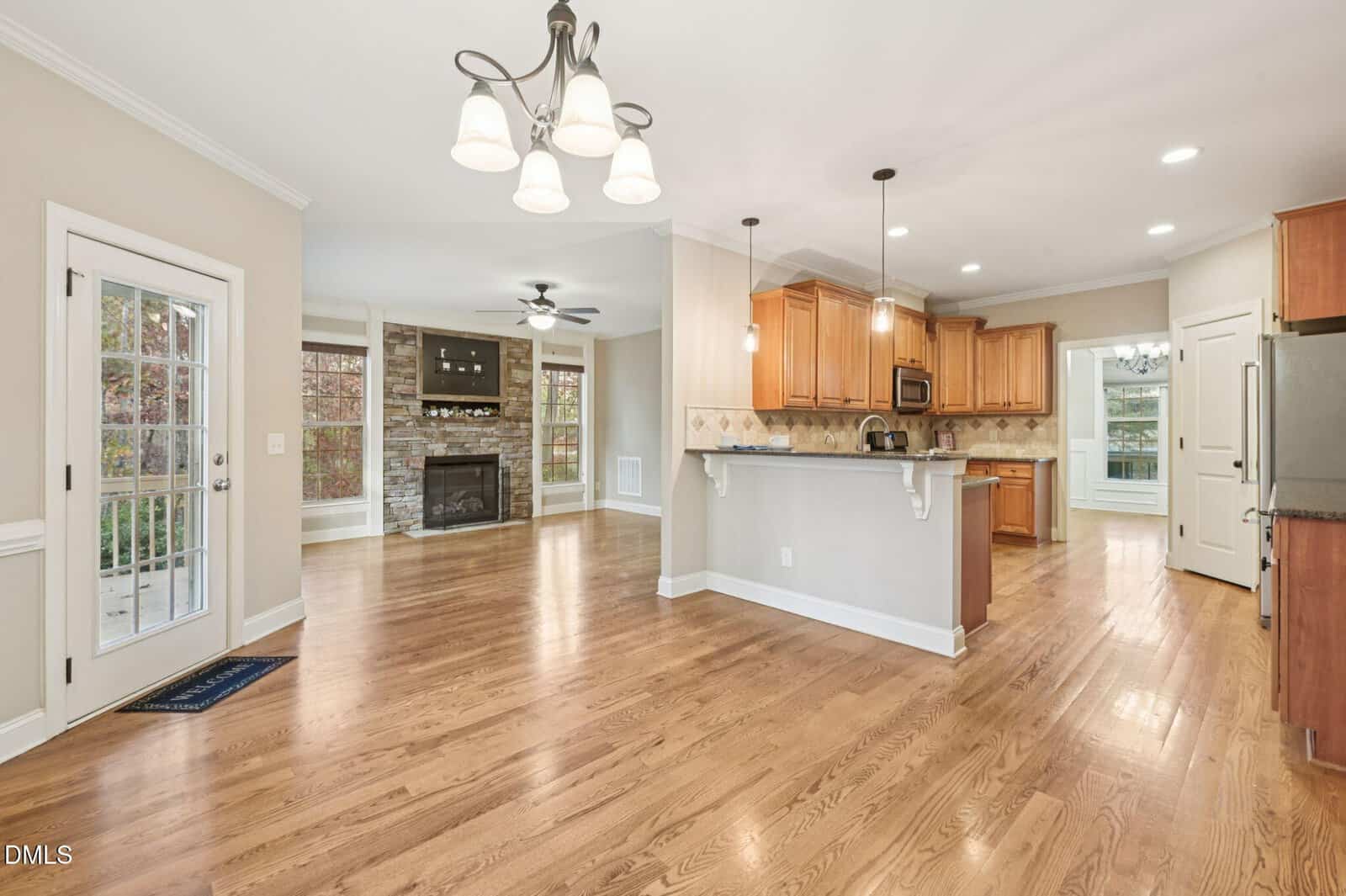 Open-concept kitchen and living area with hardwood floors, wooden cabinets, stainless steel appliances, a breakfast bar, stone fireplace, ceiling fans, large windows, and a door leading outside.