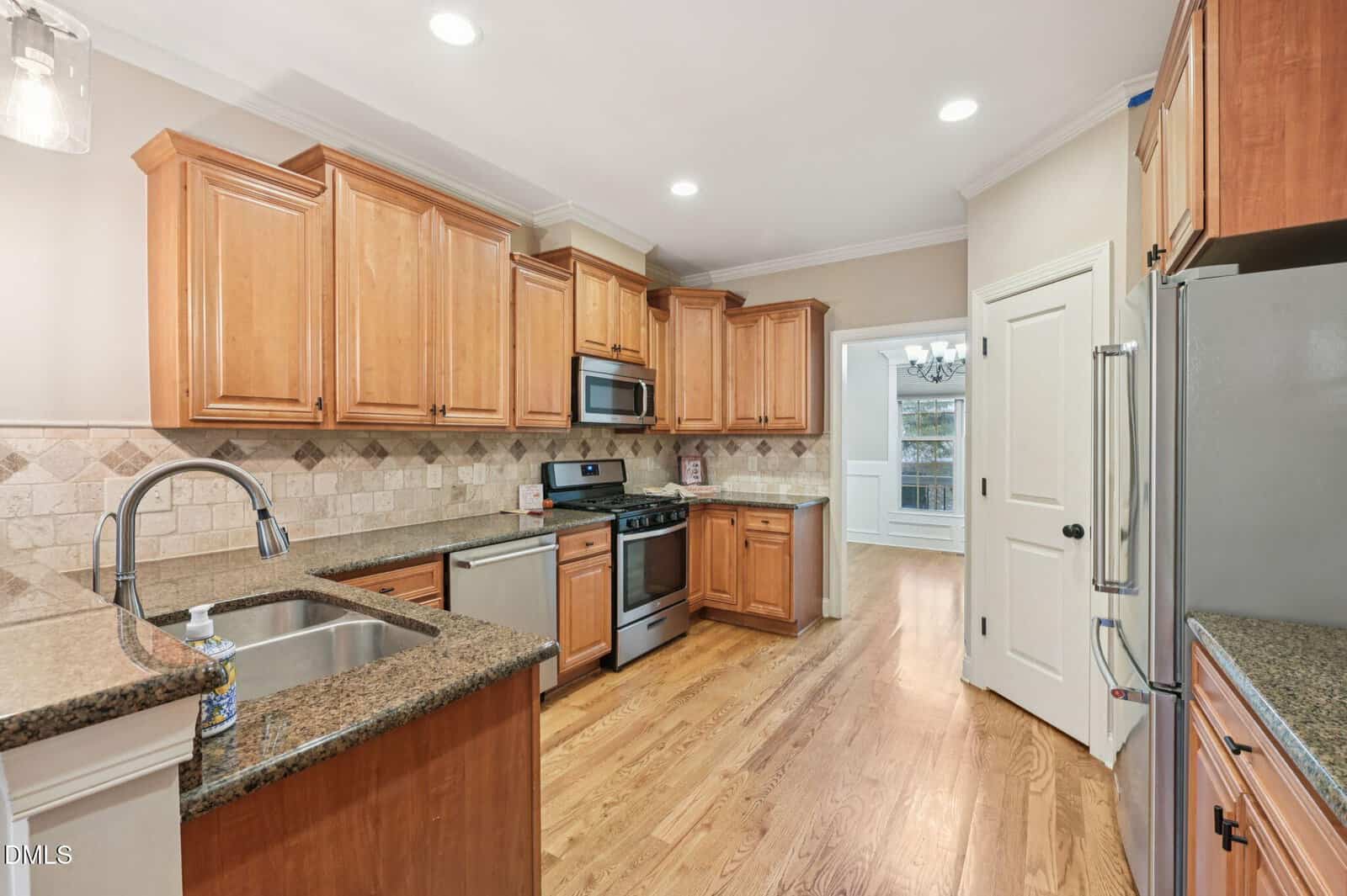 Spacious kitchen with light wood cabinets, granite countertops, stainless steel appliances, a gas stove, hardwood floors, and a tiled backsplash. An adjacent dining room is visible through a doorway.