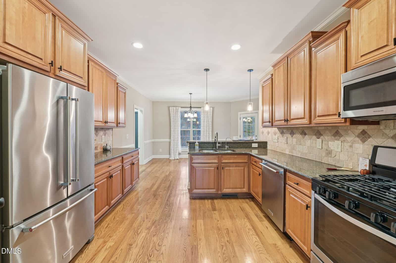 Spacious kitchen with stainless steel appliances, wooden cabinets, granite countertops, gas stove, dishwasher, and wood flooring. A dining area with windows and pendant lights is visible in the background.