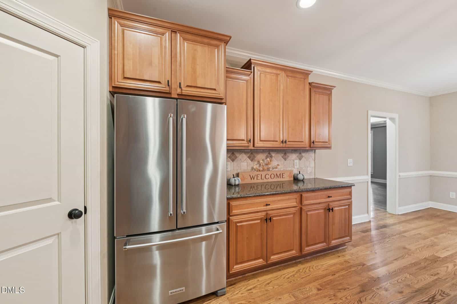 A modern kitchen with light wood cabinets, stainless steel refrigerator, hardwood floors, beige walls, and a decorative "WELCOME" sign on the counter beneath upper cabinets.