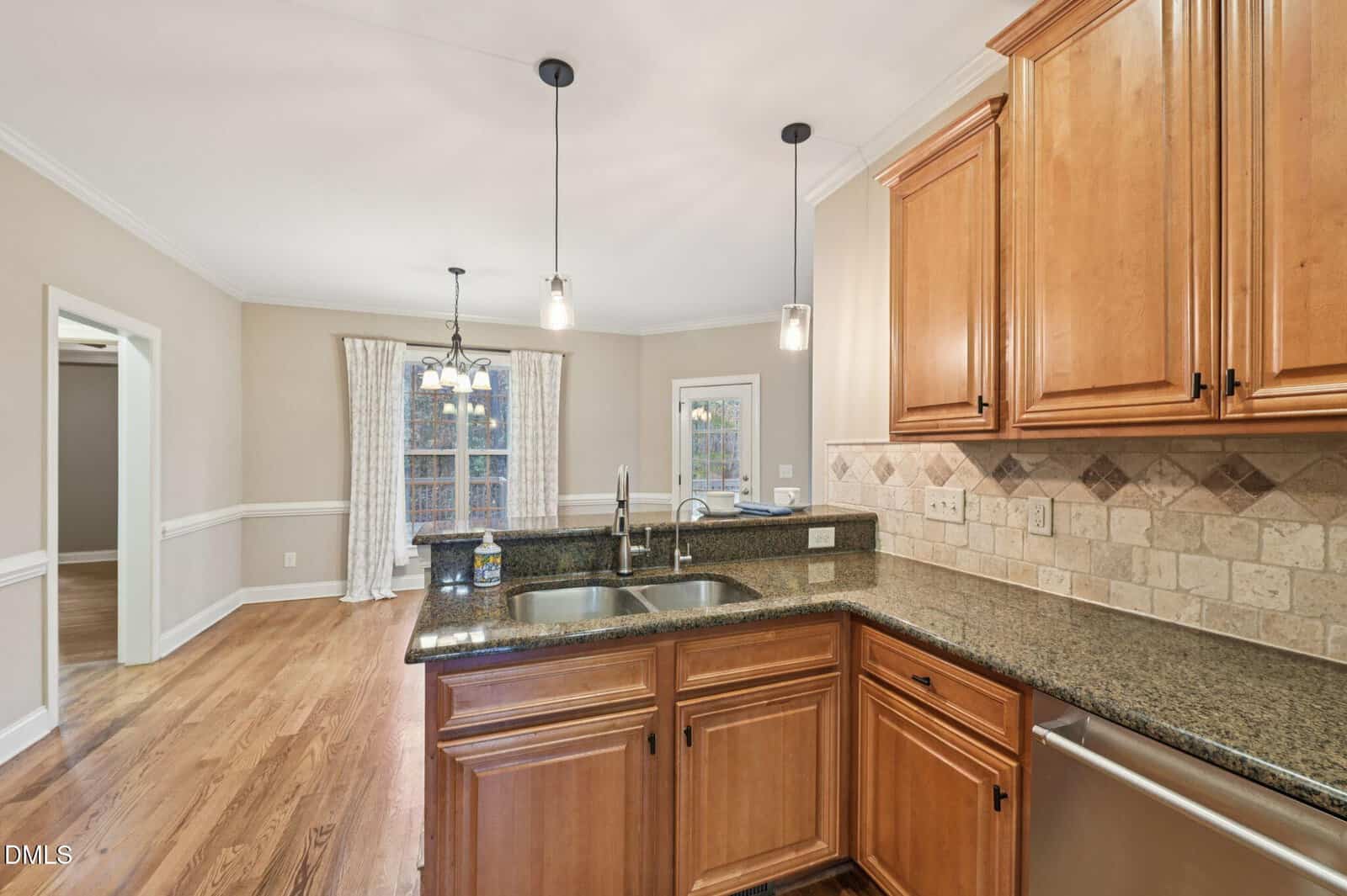 A spacious kitchen with wood cabinets, granite countertops, a double sink on an island, stainless steel appliances, tile backsplash, and pendant lights. Hardwood floors extend into a dining area with large windows and white curtains.